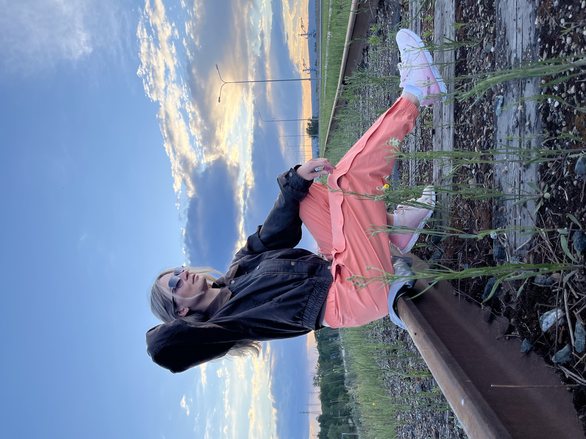 A young adult female is reclining on a railway track near Chaikovsky, Russia. She is wearing sunglasses, a dark jacket, pink trousers, and pink and white sneakers. She is resting her hands behind her head, looking upward toward a partially cloudy sky. The railway is surrounded by tall grass and other vegetation. Street lights and industrial structures are visible in the background.