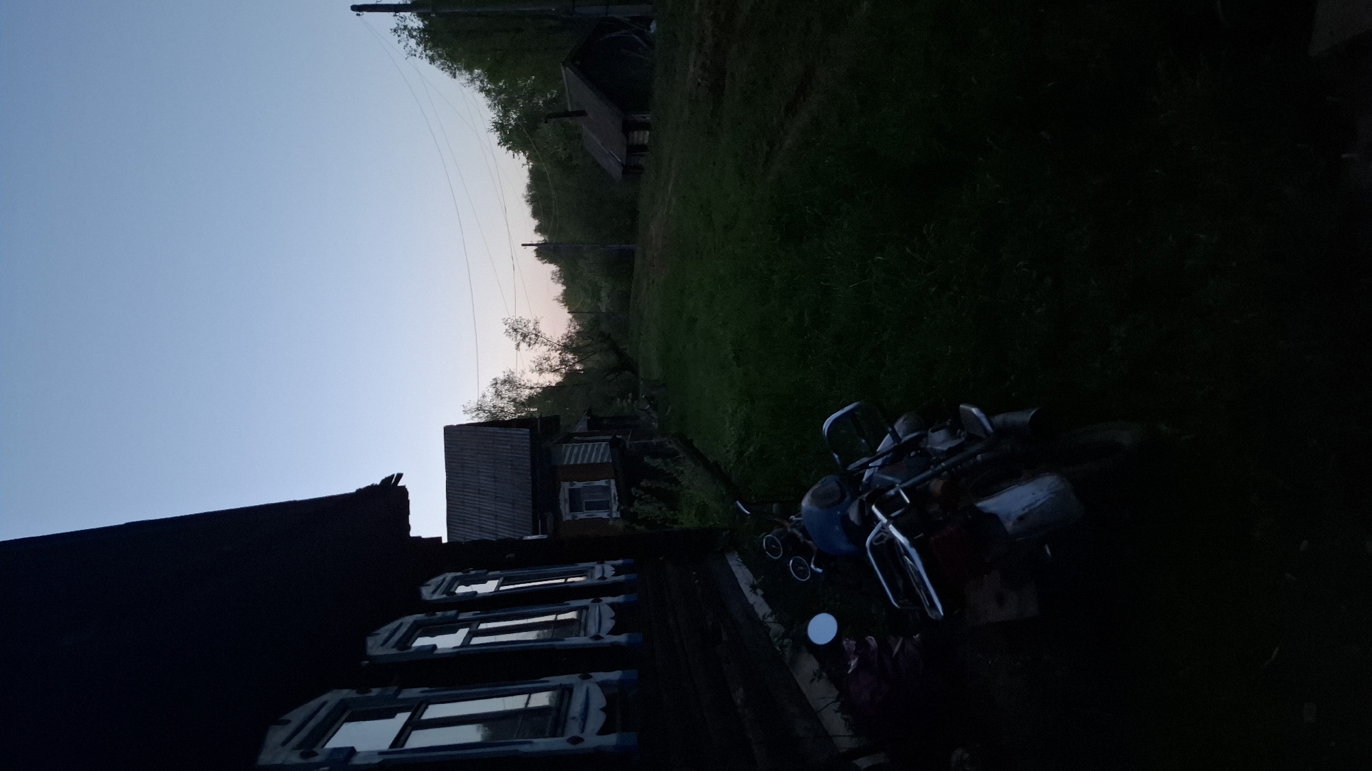 The image depicts a rural scene in the city of Шешурга, Russia, likely at dusk or dawn. A dark wooden building with visible windows occupies the lower left. In the foreground, a lawn mower rests on the grassy ground. In the background, a smaller wooden structure is visible amidst trees and vegetation. Power lines stretch across the upper part of the image against the light blue sky. The scene is dimly lit, with shadows obscuring many details.