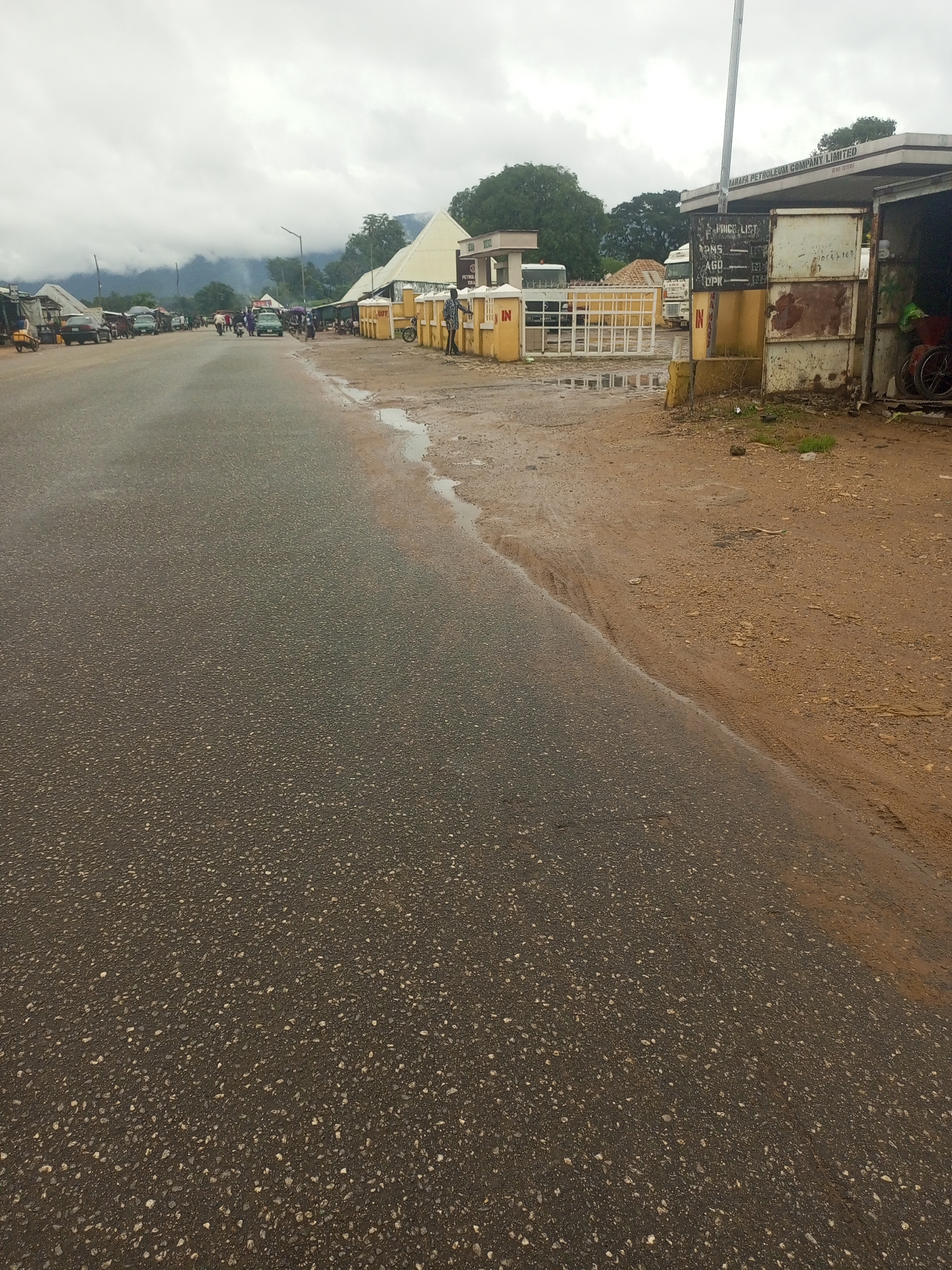 The image depicts a street scene in Serti, Nigeria, on a cloudy day. The asphalt road occupies the foreground, with a dirt shoulder to the right. Several vehicles, including cars and motorcycles, are visible on the road, some parked. On the right side of the road, there is a structure with a sign that reads "ZARAFA PETROLEUM COMPANY LIMITED". Adjacent to this is a gated entrance with a yellow wall. People are present in the scene, some walking or standing near the buildings. The overall environment suggests a commercial or transportation hub.