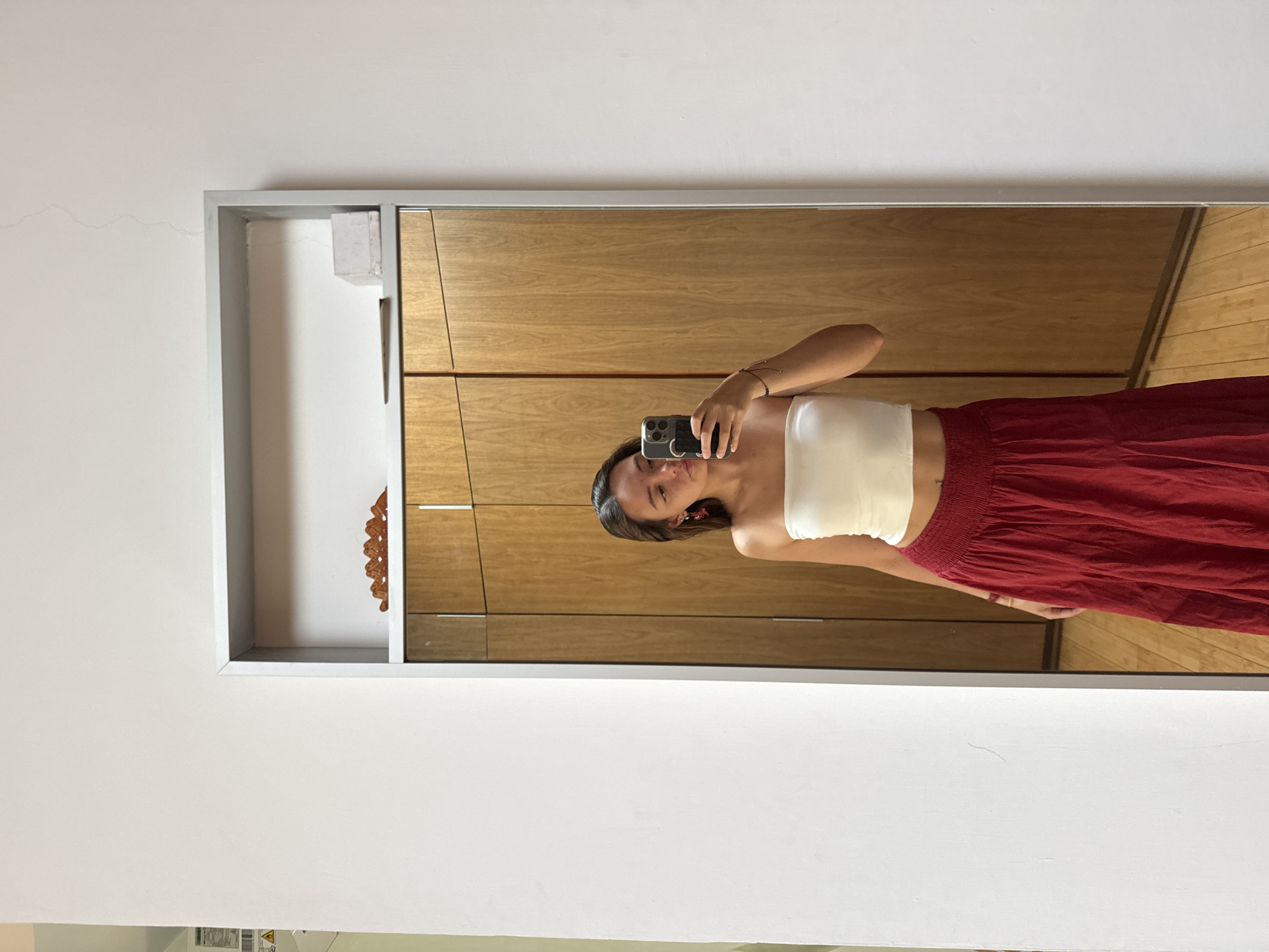 A young woman is taking a mirror selfie inside a room in what appears to be San Bernardino Tlaxcalancingo, Mexico. She is holding a smartphone in her right hand, positioned to capture her reflection. She is wearing a white tube top and a red maxi skirt. The mirror is framed with a gray border, and behind her in the reflection, there is a wooden cabinet or wardrobe, and visible portions of a wooden floor. To the left of the mirror, there's a white wall with visible cracks near the top. Within the mirror frame, there is also a decorative, possibly wooden, object visible on a shelf. The image is taken indoors, likely during daylight hours.