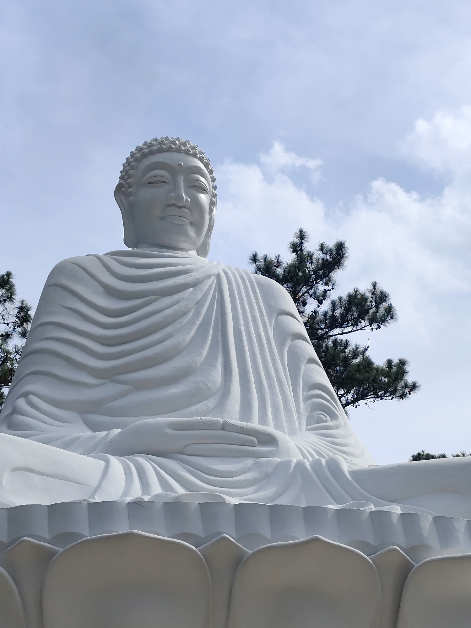 A large, white Buddha statue in a seated pose dominates the frame. The statue features a serene expression and is draped in flowing robes. The base of the statue appears to be a stylized lotus flower. Behind the statue, green pine trees partially obscure a sky with scattered clouds. The statue is located in Da Lat, Vietnam.