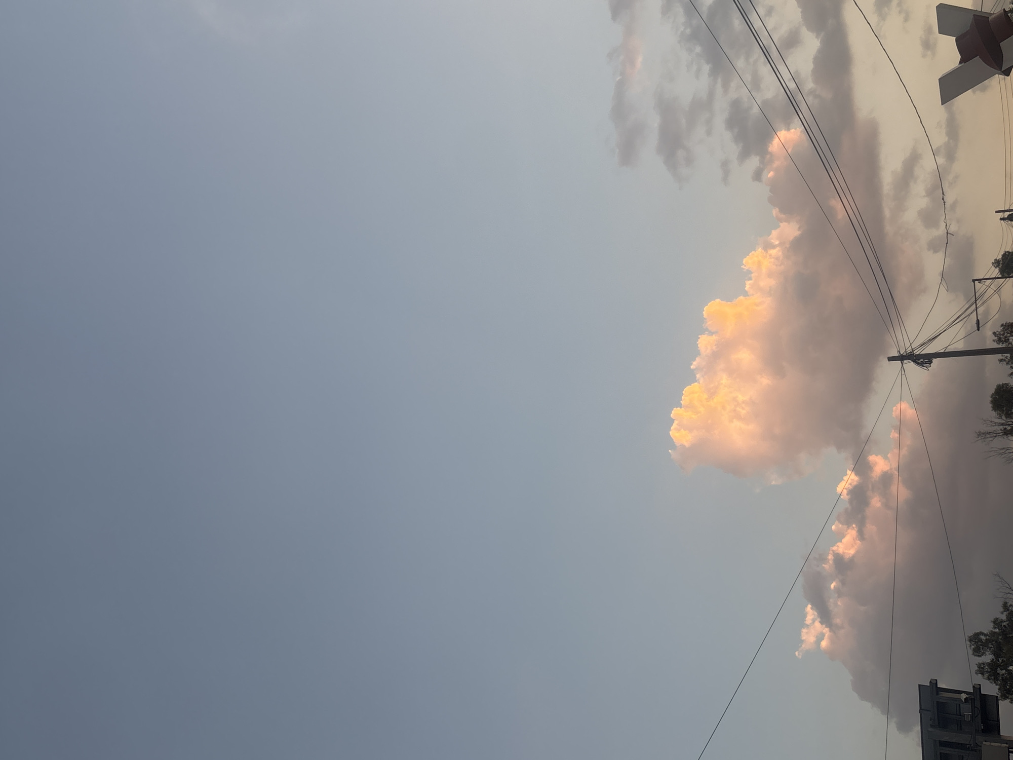 The image shows a sky with light gray-blue tint, filled with puffy clouds that appear to be illuminated by the setting sun, giving them an orange glow. Power lines traverse the upper right portion of the image. Trees and a building are also visible in the lower right corner. A railroad crossing signal is in the upper right corner. The location is Buenavista, Mexico.