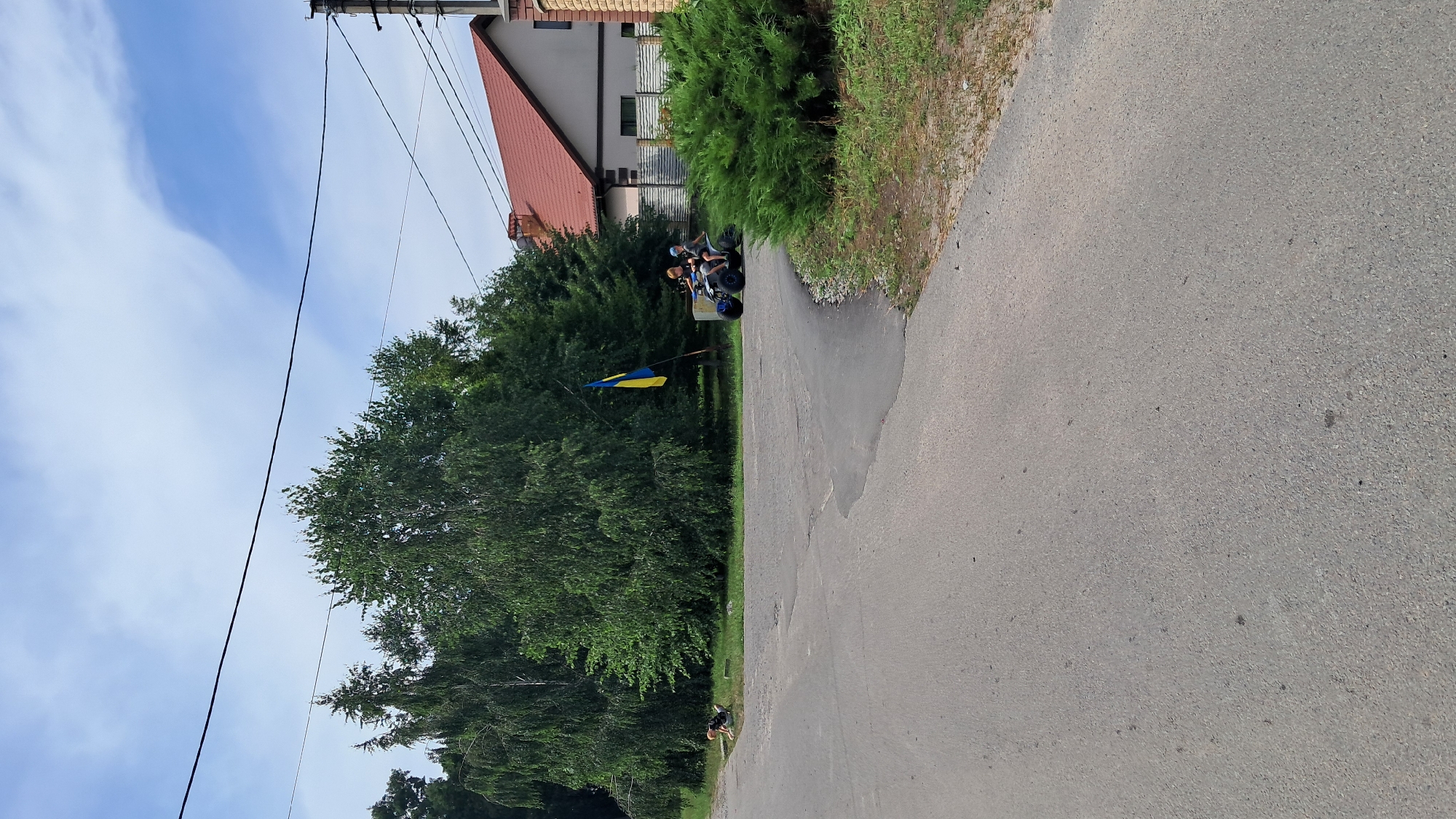 In Kryvushi, Ukraine, a two-lane asphalt road curves to the right, bordered by grass and trees. On the left side of the road, a person is lying on the ground near a patch of grass. On the right side of the road, two people are riding an ATV. Behind them, a Ukrainian flag is flying from a pole. A residential building with a red roof and a brick chimney is visible in the background, along with a section of a brick wall and a dense green hedge. The sky above is a mix of blue with scattered white clouds. Power lines are visible overhead.