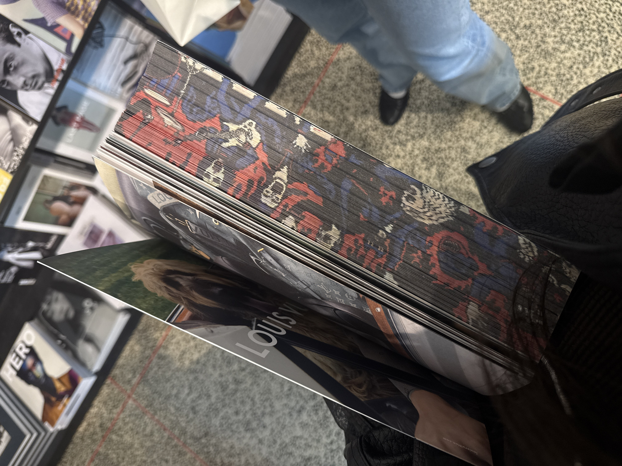 The image presents an overhead, slightly angled view of a large, thick book held open, prominently featuring its unique, intricately decorated page edges. The edges of all the book's pages are dyed with a multi-colored graphic pattern, primarily in black, red, and light blue/white. This pattern depicts stylized, abstract figures that appear to be skull-like faces, other abstract shapes, and potentially comic-book-inspired characters, creating a bold visual effect along the entire binding.
On the visible open page, the white text "LOUIS VUITTON" is clearly discernible, alongside an image that includes a person with long, wavy blonde hair. Another page shows a repeating pattern resembling the classic Louis Vuitton monogram. In the background to the left, several other books and magazines are displayed; one vertical spine is marked with "HERO", and another cover features a black-and-white portrait of a man.
In the upper right portion of the frame, a person wearing light blue jeans and black shoes is standing on a grey, speckled carpet with subtle red lines, suggesting an indoor public or commercial setting such as a bookstore, gallery, or exhibition in London. A black, textured leather item, possibly a bag or jacket, is visible in the bottom right foreground. The image is captured from a perspective looking down at the book, implying it is being held or closely examined.