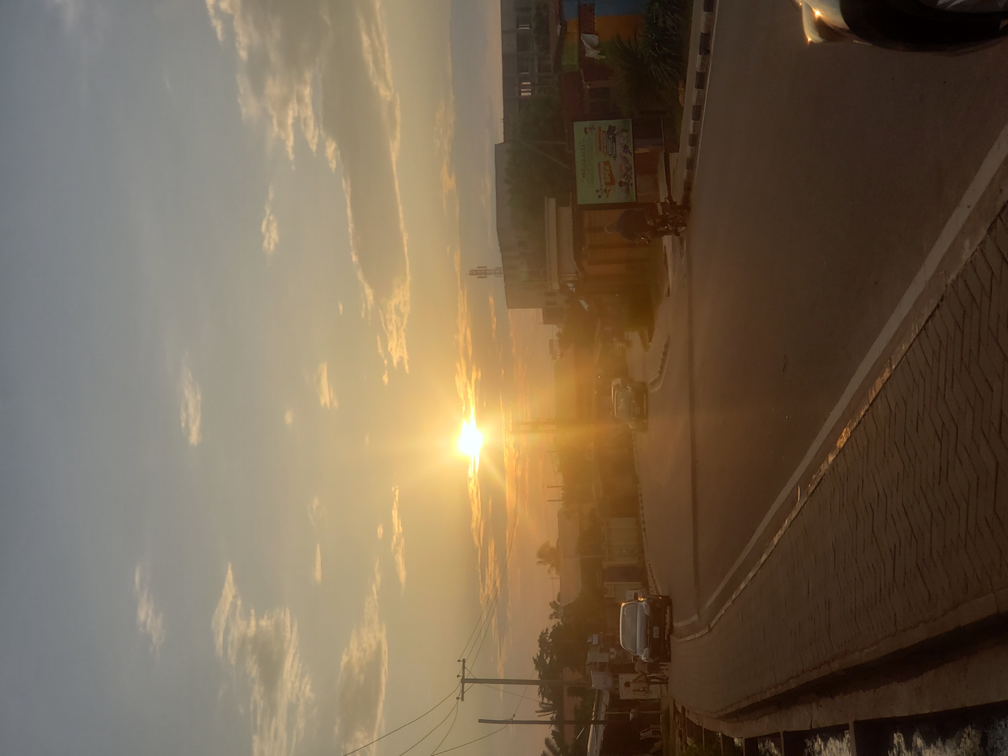 The image captures a street scene in Narayi, Nigeria, at sunset. The sun, a bright orb, is positioned in the upper center of the frame, casting a warm, golden light across the sky and the surroundings. The sky is filled with scattered clouds, tinged with orange and yellow hues from the setting sun.
In the foreground, a paved road curves to the left, with a brick-lined walkway on the left edge. Several vehicles are present on the road, including a dark sedan approaching from the left and another vehicle further down the road. A person is seen riding a motorcycle on the right side of the road, and a few pedestrians are visible near the left side of the road.
On the right side of the street, there are buildings and structures, including a billboard with some text and images. Power lines and poles are visible on the left side of the scene. The overall atmosphere is serene, typical of a late afternoon or early evening in a residential or commercial area.
