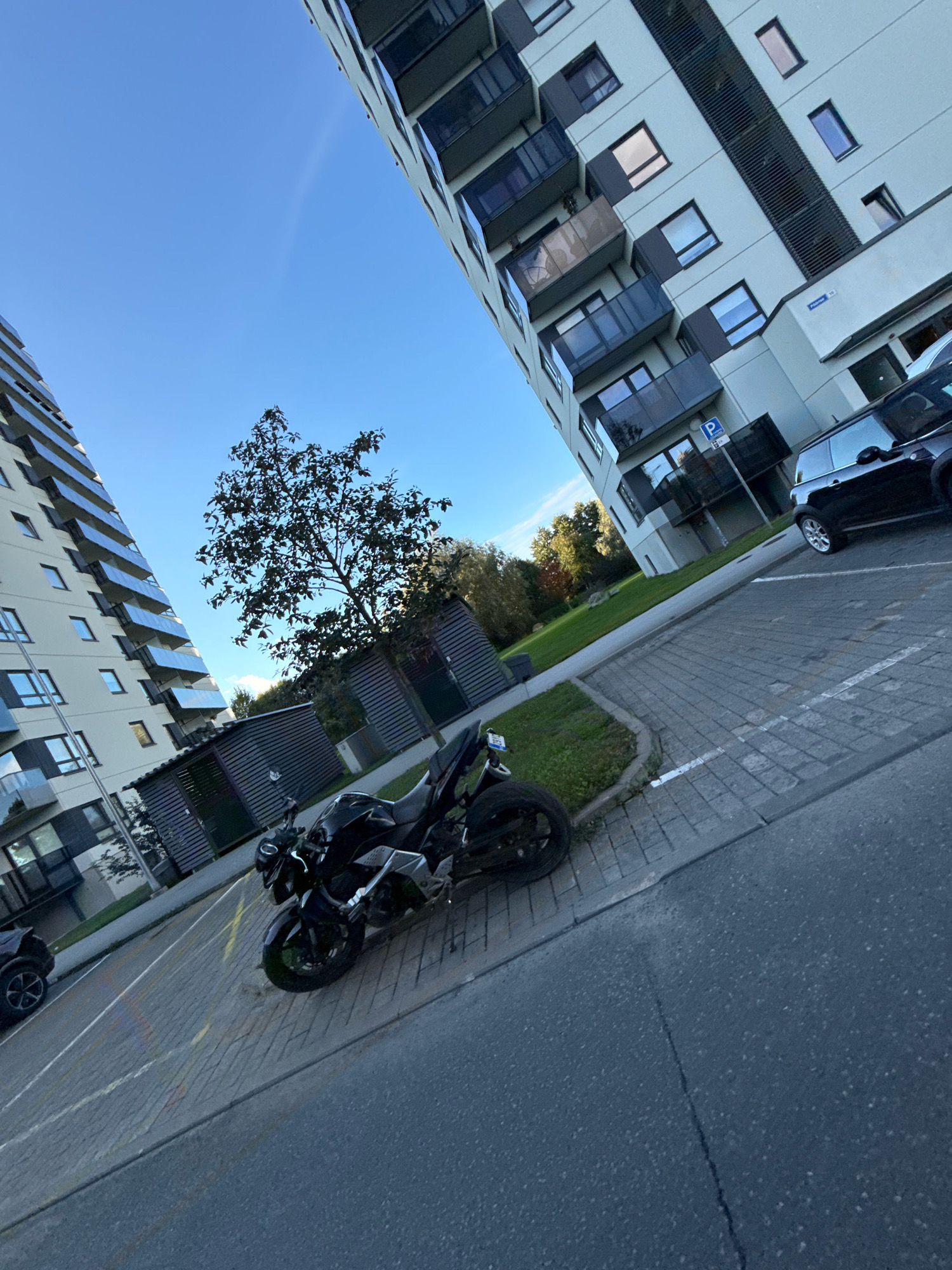 In the city of Tallinn, Estonia, a black motorcycle is parked on a paved surface in front of a modern, multi-story apartment building. The building features a light-colored facade with numerous balconies and windows. To the left of the frame, another similar apartment building is partially visible. A black car is parked further down the paved area near the first building, next to a sign indicating parking. A tree with sparse foliage stands between the motorcycle and the buildings, and beyond the tree, more green space and trees are visible. The sky is clear and blue, suggesting a daytime scene. The image is captured from a low angle, emphasizing the scale of the buildings and the motorcycle.