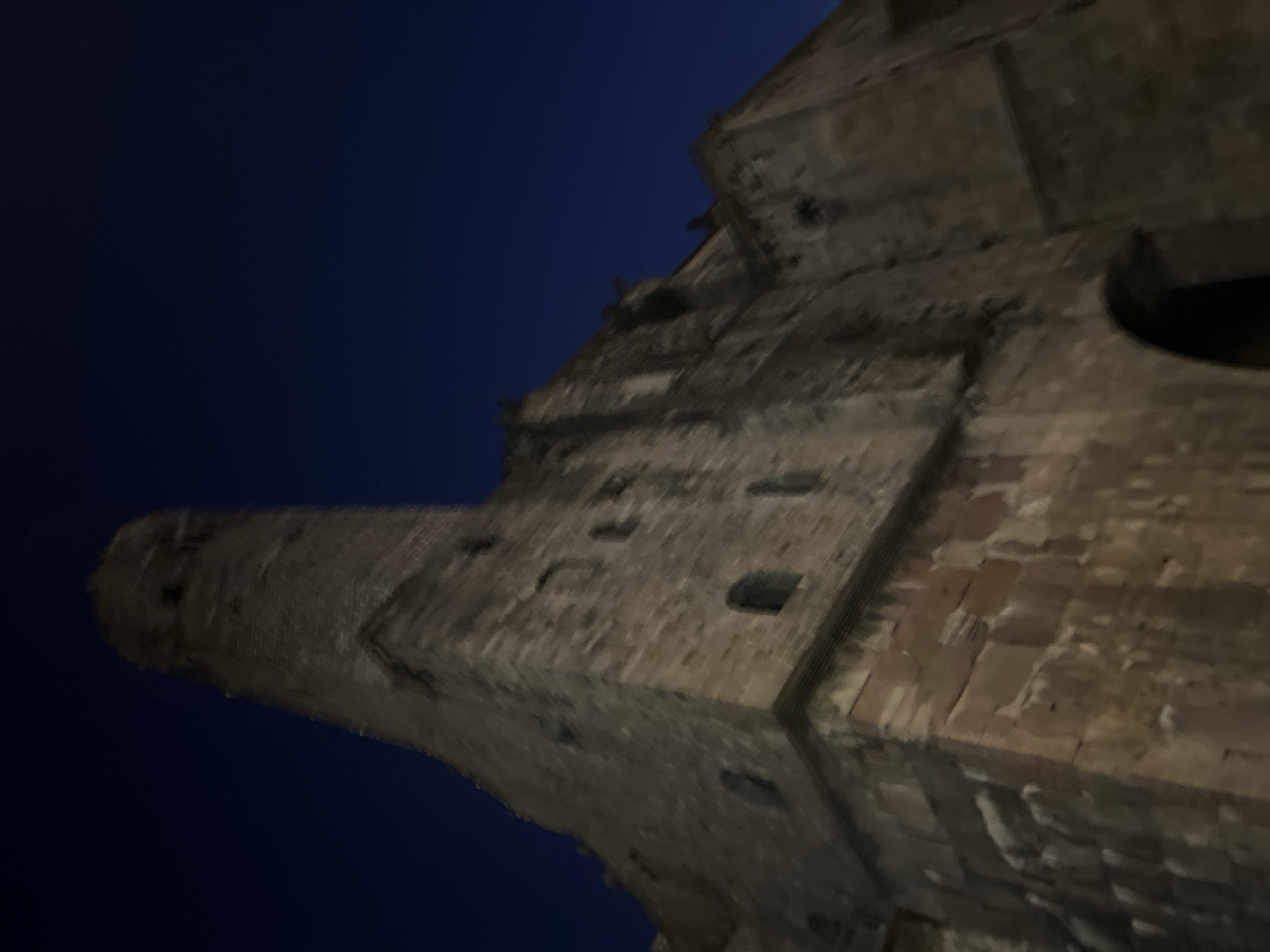 The image shows a low-angle, upward view of a stone tower and part of a building at night. The structure appears to be made of rough-hewn stone blocks, with visible mortar. Small, dark openings, likely windows, are present on the tower and the building's facade. The sky is a deep, dark blue, indicating it is nighttime. The lighting suggests artificial illumination, casting shadows on the stone surfaces. The building's architecture suggests it could be historical, possibly a church or a medieval tower. Based on the architectural style and the presence of similar structures in the city, this is likely the bell tower of the Basílica de Santa Maria del Mar in the El Born district of Barcelona, Spain. No people or vehicles are visible in the frame.