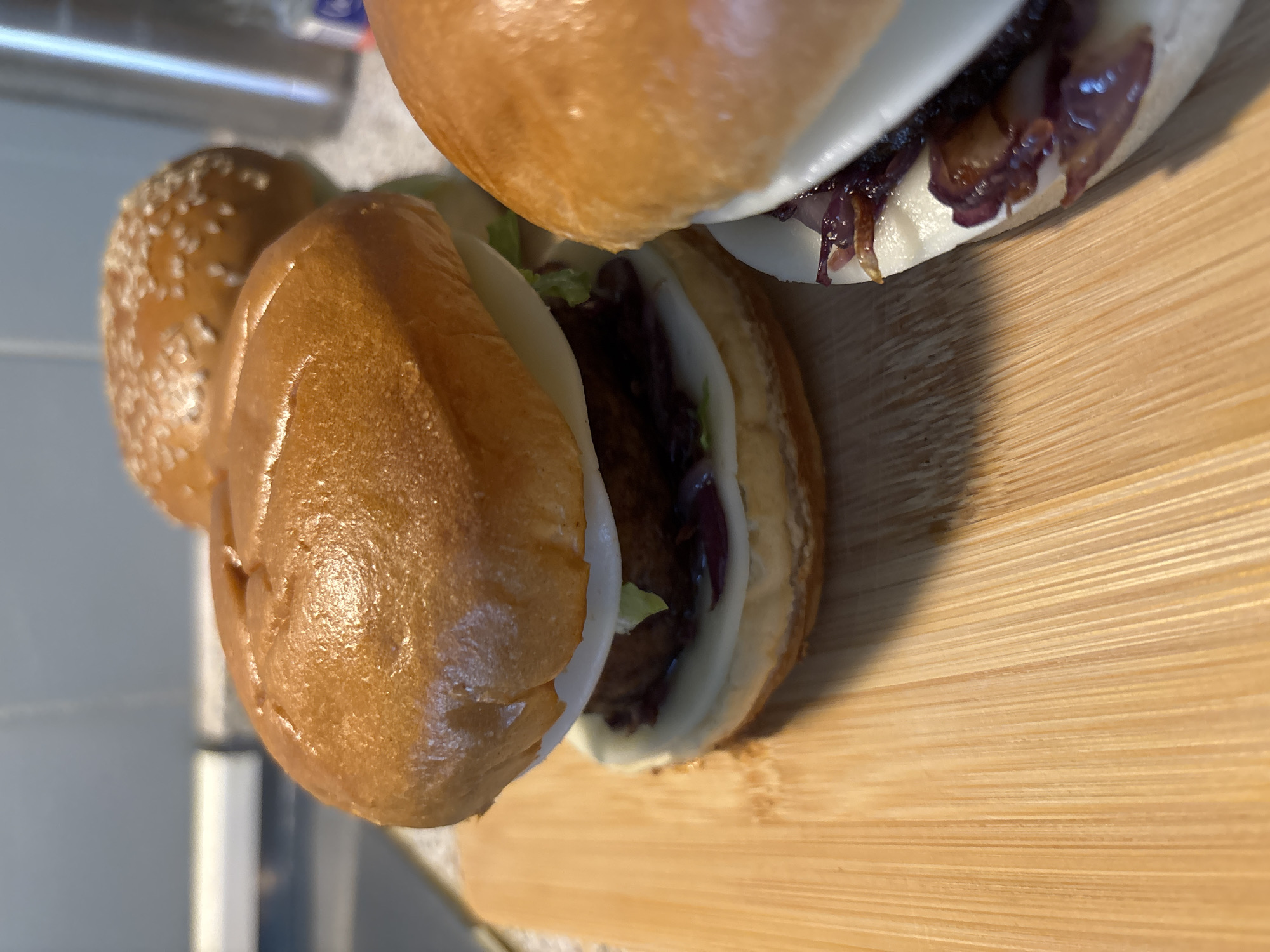 The image shows three hamburgers on a wooden cutting board. The hamburgers have golden brown buns, white cheese, a dark patty, and red onion. One hamburger is in the foreground, to the left, and is mostly in focus. Another hamburger is behind it, to the right, and is also mostly in focus. A third hamburger, with sesame seeds on the bun, is in the background and is out of focus. The cutting board is made of light-colored wood with parallel grain. The background shows a gray tiled wall and some metallic objects. The scene appears to be in a kitchen. The image does not provide any information about the city of Wörth am Main, Germany.