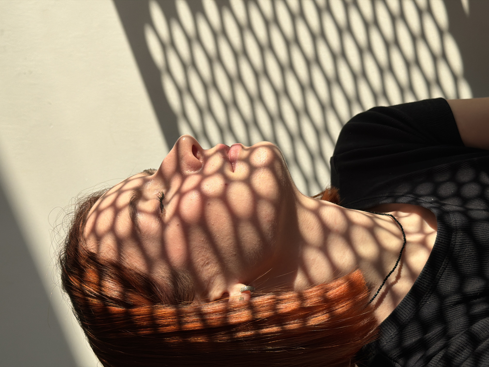 A female subject is depicted in a close-up profile view, from the upper chest upwards, against an unadorned, light-colored wall. Her eyes are closed, and her head is angled slightly upwards and to the right, oriented towards a strong, directional natural light source. She has reddish-orange, bob-length hair. A silver-colored hoop earring is visible in her left earlobe, with a smaller stud earring positioned higher on the lobe. A thin, black beaded choker necklace encircles her neck. She is wearing a black, short-sleeved, ribbed garment. A distinct hexagonal grid shadow pattern is cast across her face, hair, and clothing, extending onto the background wall. This scene is situated within a building in the city of Chaikovsky, Russia.