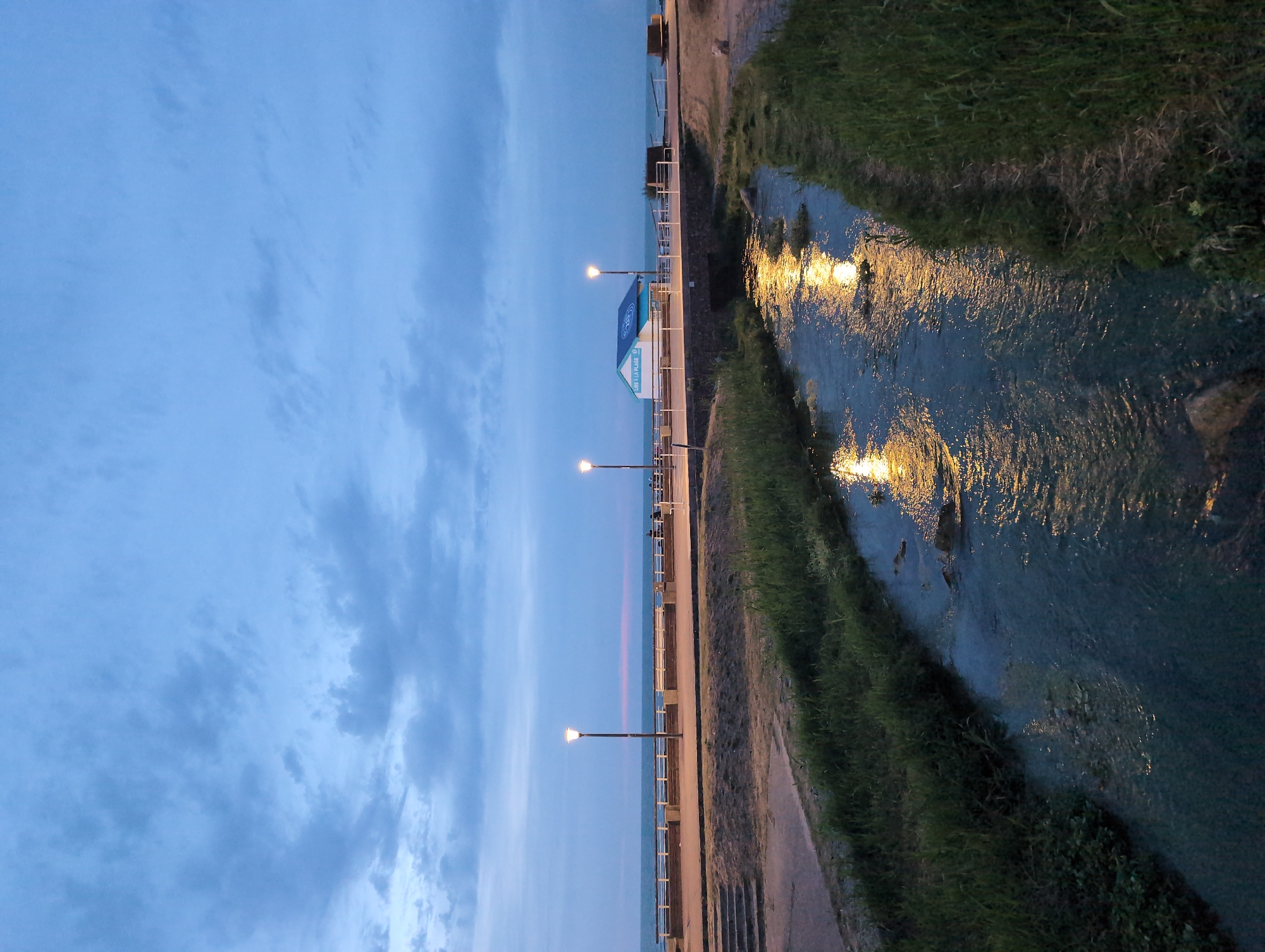 The image depicts a tranquil evening scene in Veules-les-Roses, France. A small, clear stream flows through the foreground, its surface reflecting the warm glow of streetlights. Lush green grass lines the banks of the stream. In the background, a promenade runs alongside a body of water, possibly the English Channel. A few scattered streetlights illuminate the promenade, and a small, blue and white building with text "VIE À LA PLAGE" is visible. The sky is a deep blue with scattered clouds, suggesting either dusk or early dawn. A few indistinct figures can be seen on the promenade in the distance. The overall atmosphere is peaceful and serene, capturing the quiet beauty of the seaside town.