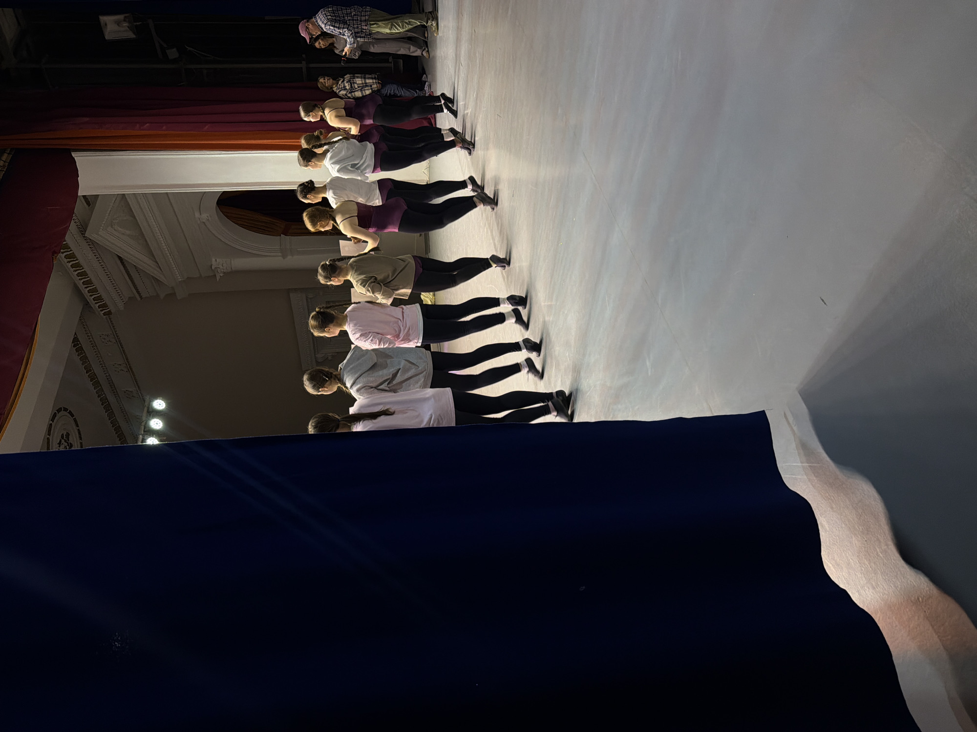 A straight line of ten young women in sportswear is standing on a stage in Perm, Russia, during a dress rehearsal or practice session, with two men observing in the background. The women are facing away from the camera. They are wearing various colored shirts: some are pink, white, gray, or purple, and black leggings. The floor of the stage is a light gray. A section of draped black fabric and a light gray curved object are in the foreground. Part of the building's architecture, including decorative columns and ceiling fixtures, is visible in the background, to the left. The men are wearing plaid shirts.