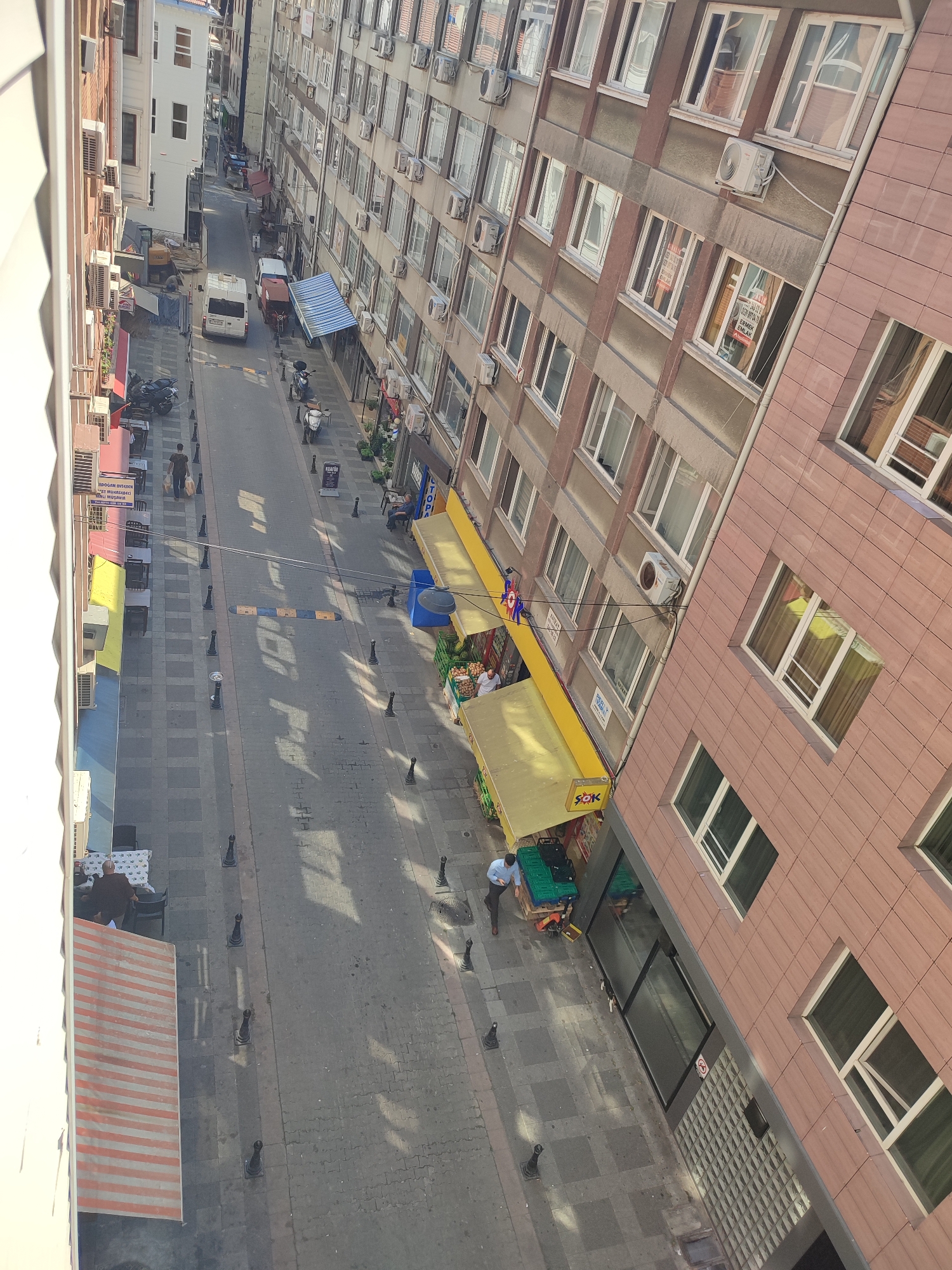 The image captures a street scene in Kadıköy, Turkey, looking down from a high vantage point. The street is paved with gray interlocking blocks and lined with buildings. On the left, a white building with an awning stands adjacent to a taller, older building with multiple windows and air conditioning units. Further down the street, a white van and a red cart are parked. On the right side of the street, a modern building with a pinkish-brown tiled facade dominates, with many windows featuring air conditioning units and some with "For Rent" signs visible. A yellow awning covers a storefront below. The street is relatively narrow, with traffic calming measures like speed bumps visible. Several black bollards line the sidewalks. People are present: one individual walks down the street carrying bags, another sits at a table outside a cafe, and a person in a blue shirt is interacting with goods on a pallet near a storefront. The overall impression is of an urban environment with commercial activity and residential buildings.