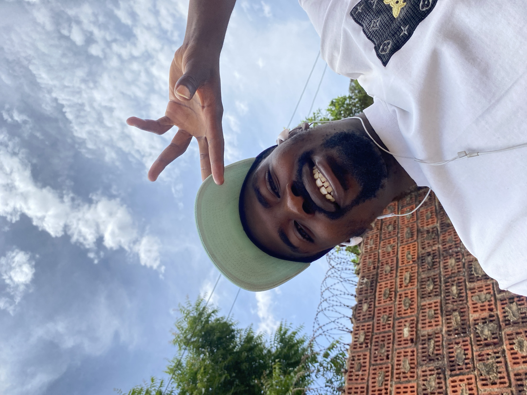 This is a low-angle, eye-level selfie-style photograph of a young man, captured outdoors under a bright, cloudy sky. The image is rotated approximately 90 degrees counter-clockwise, giving the impression that the man is looking down into the camera with his head on the right side of the frame and his hand extending from the top.
The central subject is a man with dark skin and a beard, smiling widely and showing his teeth. He is wearing a light green baseball cap and a white short-sleeved T-shirt. White wired earphones or earbuds are visible in his ears. His right hand is extended towards the camera, fingers spread in a casual gesture.
His white T-shirt features a prominent square patch on the left chest area. This patch is dark-colored (possibly black) and is embroidered with golden designs, including stylized "LV" initials, stars, and what appears to be a small cartoon bear or similar animal.
The background primarily shows a bright blue sky with numerous scattered white clouds. On the right side, a distinctive brick wall structure is visible, characterized by rows of square hollows or ventilation blocks, topped with barbed wire. Green foliage from trees or bushes can be seen behind the man's head and extending into the middle-left background.
The photograph conveys a cheerful and casual mood, taken during bright daytime conditions. The unique brick wall design and the general setting align with the provided location context of Jos, Nigeria. No explicit text is clearly legible beyond the stylized initials on the T-shirt design.