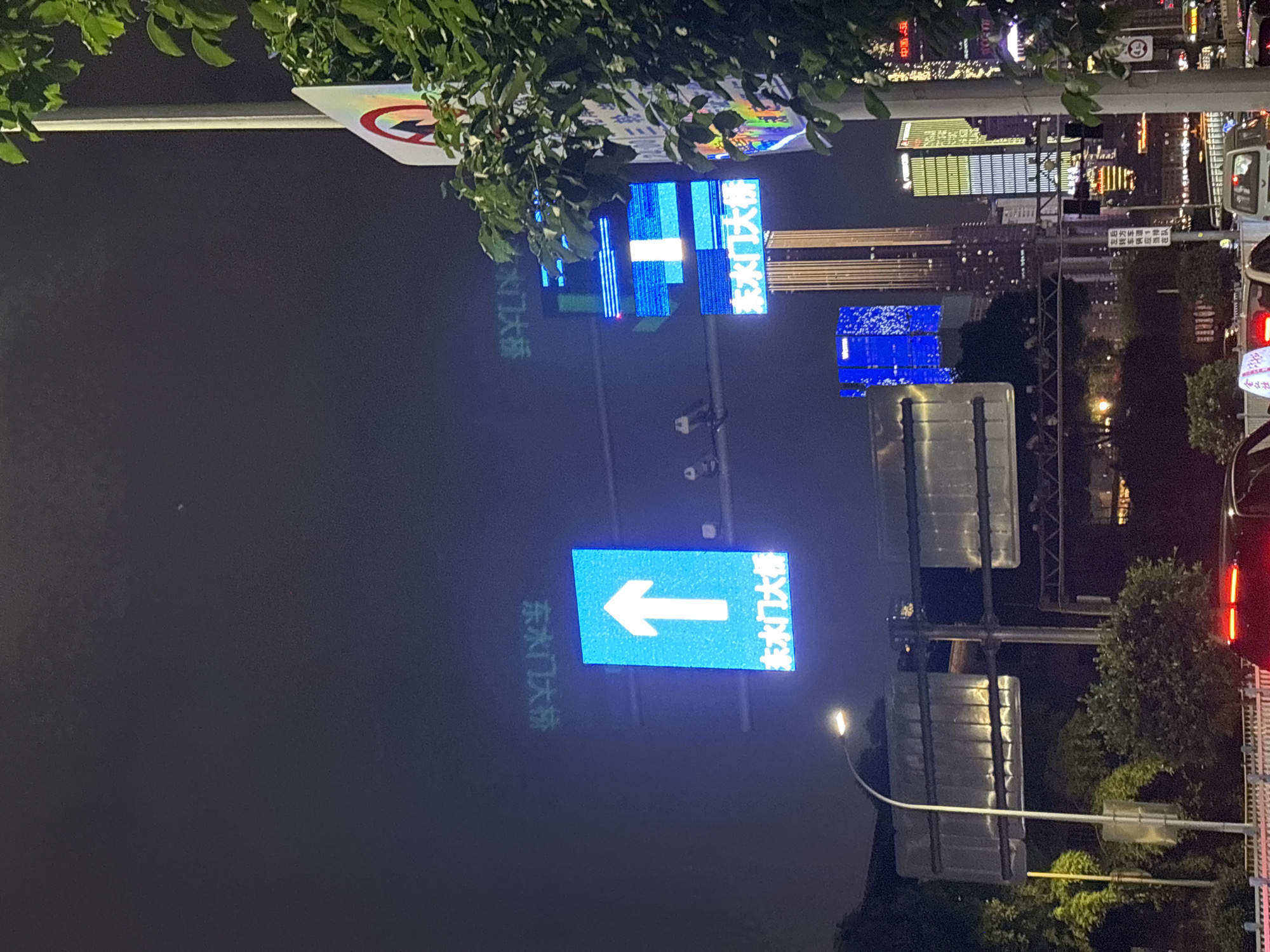 This is an exterior night shot in Nan'an District, China. It features illuminated traffic signs, with one pointing to the left and another indicating "Dongshui Gate Bridge" (东水门大桥) in Chinese characters. The signage is positioned on poles along a street. The background reveals buildings and city lights. There are trees in the foreground and the right side of the image. Vehicles with lit tail lights are present on the road. There is also a partially visible sign in Chinese mentioning "Left turn vehicles" and "queuing."