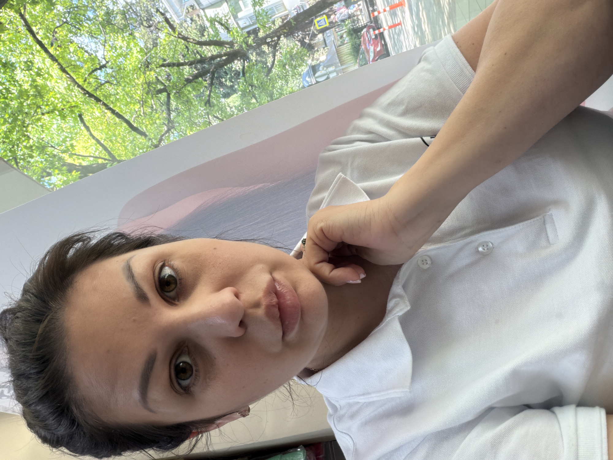 A woman is taking a selfie. She has brown hair pulled back, brown eyes, and is wearing a white polo shirt. Her left hand is resting on her chin. She appears to be indoors, with a large poster or banner behind her and a window to her right showing trees and a street scene with cars and pedestrians. The location is the city of Sochi, Russia.
