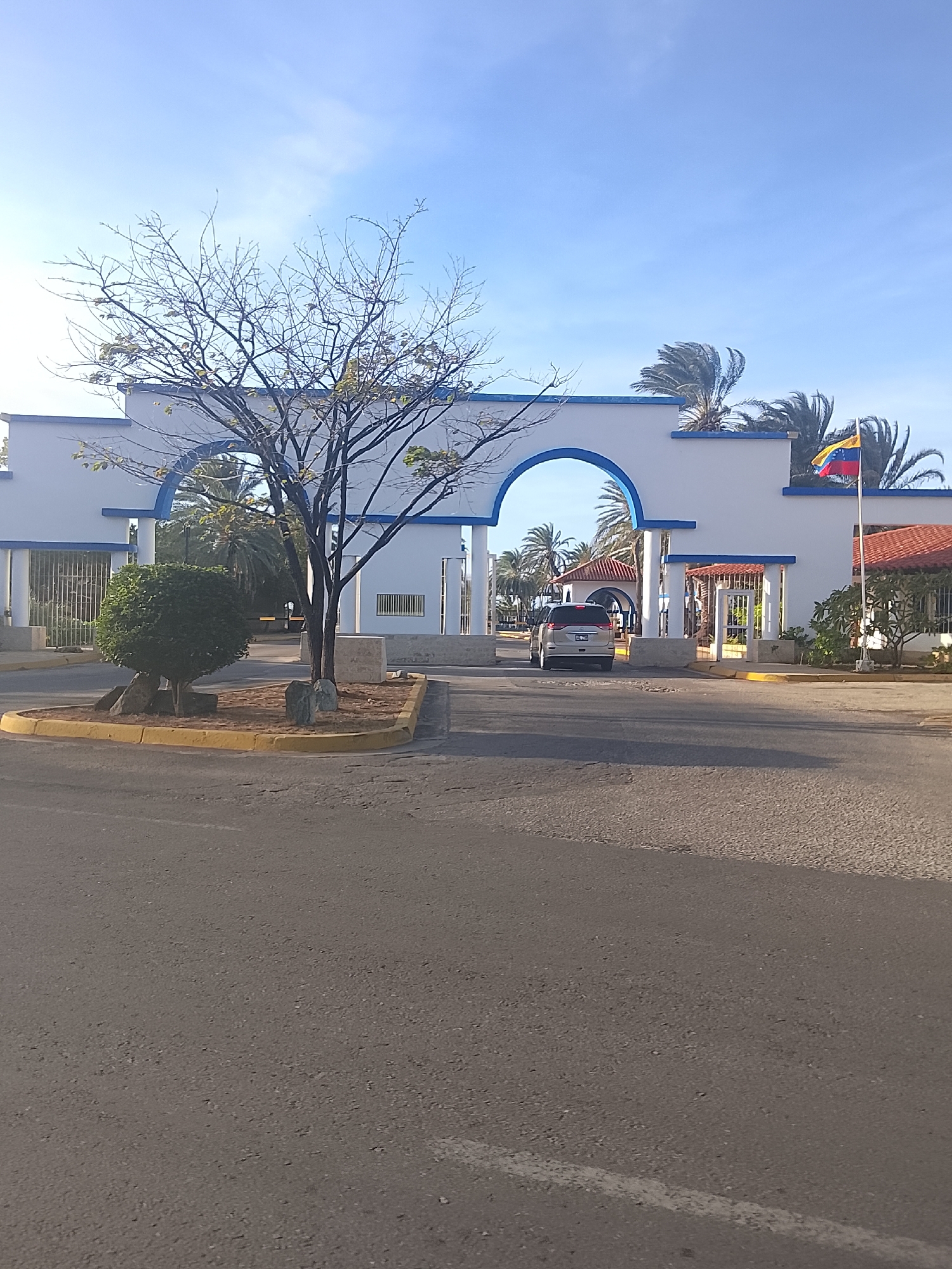 The image captures an entrance in Altagracia, Venezuela. A white archway, trimmed with blue, serves as the main gateway. A Venezuelan flag is to the right of the archway. A silver minivan proceeds through the archway. There is a tree and a rounded bush in front of the archway entrance. The foreground shows an asphalt road. Palm trees can be seen behind the archway.