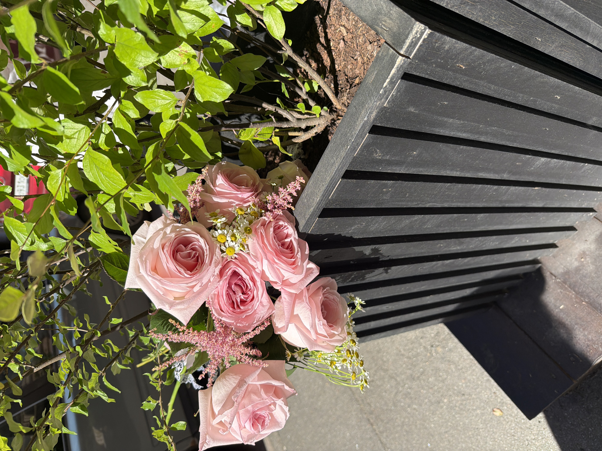 A close-up, eye-level outdoor shot shows a planter box filled with a bushy green plant and a bouquet of light pink roses and smaller pink flowers. The planter box is made of dark, vertical wooden slats. Sunlight casts shadows on the sidewalk next to the planter. The scene appears to be a business or residential area. No people are visible. The image does not provide specific information to determine its location within Saint Petersburg, Russia.