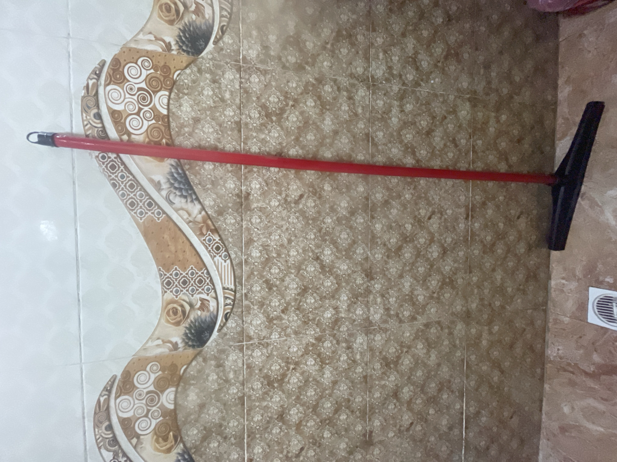 A red-handled broom with a black flat head stands upright against a tiled wall in Masazir, Azerbaijan. The wall features a decorative pattern of wavy bands with floral and geometric motifs in shades of brown, tan, and white. Below this is a tiled section with a repeating small floral pattern in a light brown hue. The floor in front of the wall is also tiled in a mottled pinkish-brown. To the left of the broom's base, a white circular grate is visible on the floor. A pink object, possibly a bag or container, is partially visible on the far right. The lighting suggests an indoor environment.