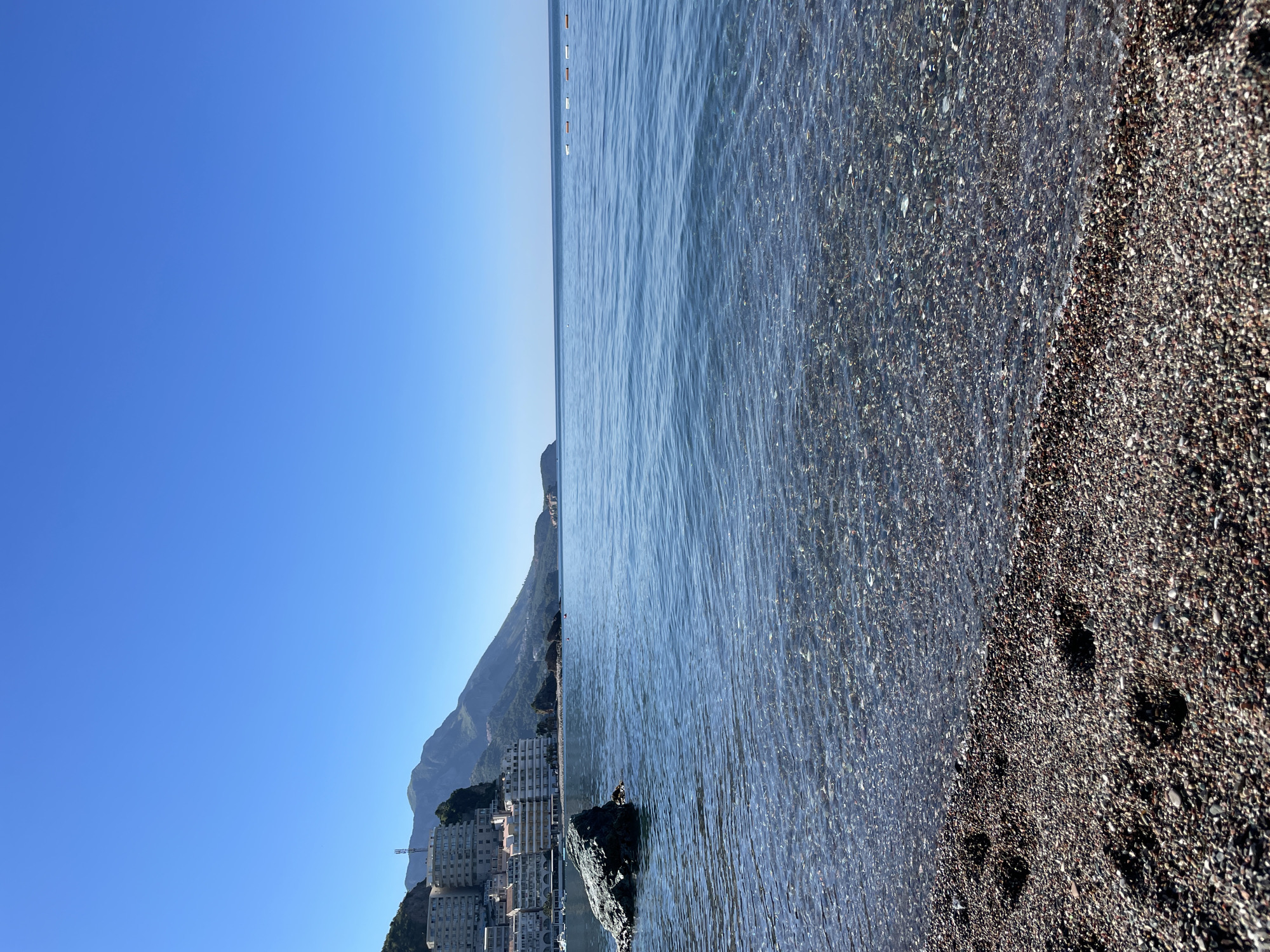 The image shows a view of the coastline in Rafailovići, Montenegro. The sky is a clear blue with no clouds. The water is clear and blue with small waves lapping onto a dark, pebbled beach. Buildings can be seen clustered at the base of mountains rising in the background along the coast. There are distant boats on the water.