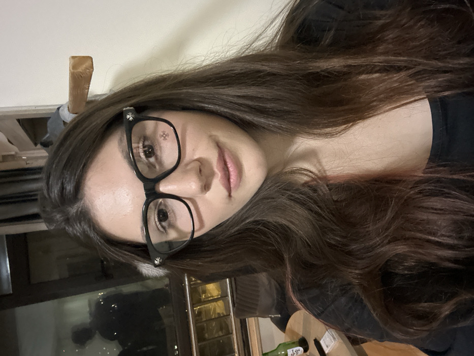 A female subject, appearing to be in her twenties or early thirties, is positioned centrally, facing forward and looking directly at the viewer. She has long, dark brown hair with subtle reddish undertones, worn down and wavy. She is wearing black-framed glasses featuring decorative cross-like emblems on the outer corners. Her expression is neutral with a slight smile. She is dressed in a dark-colored, long-sleeved top with a wide neckline. A small, white, possibly fluffy object is partially visible near her left temple.
The scene is an indoor, likely residential, setting within Huangdao District, China. To the left of the subject, a window reveals a dark exterior, indicating night, with blurred distant lights. A faint, dark reflection of a person, possibly male and wearing a hat or hood, is discernible in the window. Below the window, a small, light-colored round table is present, holding a green glass bottle (consistent with a soju or beer bottle), a dark rectangular object, and a stack of small white rectangular items. Part of a dark-colored upholstered chair or sofa is visible near the table. To the right of the subject, the background consists of a plain, light-colored wall. A wooden beam or protrusion extends from the upper right portion of this wall, with a dark, indistinct object hanging or resting above it.