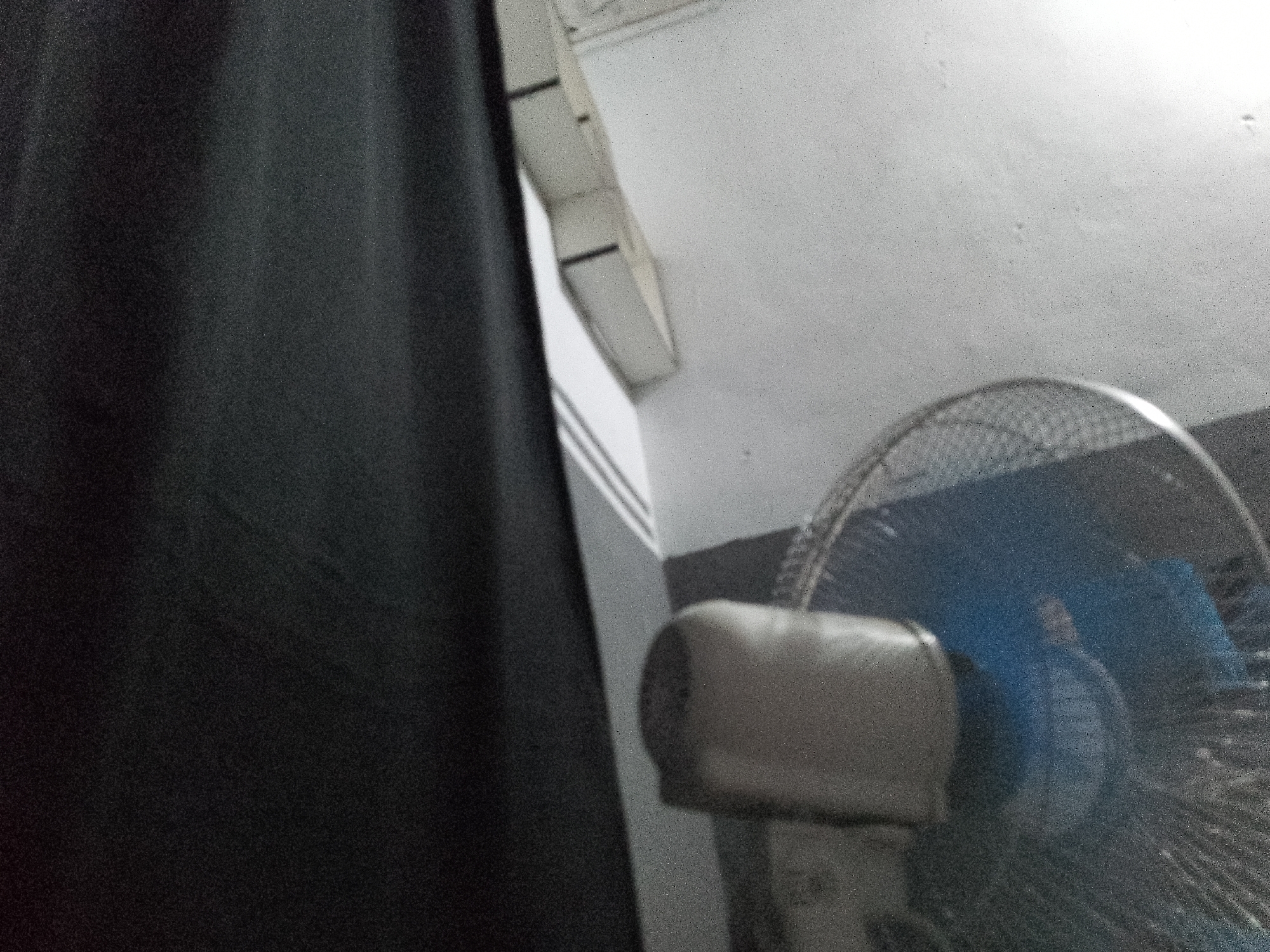 A close-up, low-angle, medium shot shows a black curtain on the left side of the frame, and a fan on the right. The fan has a silver grill and a blue motor housing. The wall behind the fan is white and textured. The scene appears to be indoors. The location in Senti, Nigeria, cannot be determined from the image.