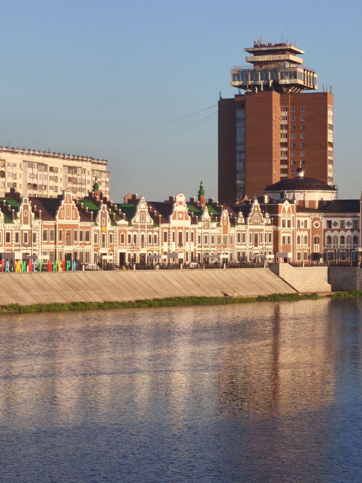 This image captures a waterside view of Yoshkar-Ola, Russia, specifically showcasing the embankment and a row of colorful buildings with distinct architectural styles. The prominent feature is a series of multi-story buildings constructed in a European-inspired design, characterized by steeply pitched roofs, decorative facades, and large windows. These buildings are brightly painted in orange, white, and green, creating a vibrant contrast against the blue sky. In the background, a taller, modern brick building with a unique, layered top structure dominates the skyline. A wide river or body of water occupies the foreground, reflecting the buildings and the warm light of what appears to be either sunrise or sunset. Along the embankment, a concrete wall separates the water from the buildings, and a section of green grass is visible at the top of the wall. Colorful three-dimensional letters spelling out a message in Russian are placed along the embankment in front of the buildings. A single person is visible walking on the embankment, near the water's edge. The overall scene conveys a sense of urban development integrated with historical architectural influences, set against a serene natural backdrop.