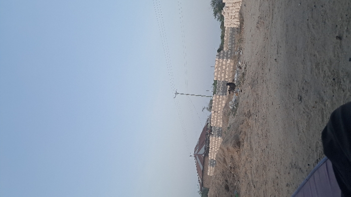 The scene depicts a rural or developing area in Walu, Kenya, under a clear, light blue sky. The ground consists of uneven, light brown sand and dirt, with several mounds of excavated earth. In the mid-ground, a single-story structure with a reddish-brown corrugated metal roof is visible on the left. Adjacent to it and extending to the right are unfinished perimeter walls constructed from light-colored concrete blocks, with additional stacks of blocks to the far right, indicating active construction. A utility pole with multiple overhead wires stands centrally in the mid-ground. A dark-feathered bird, possibly a chicken, stands on the sandy ground in front of the unfinished walls, to the right of the center. In the bottom-left foreground, a sliver of a purple object and dark fabric are partially visible. No human figures are present.