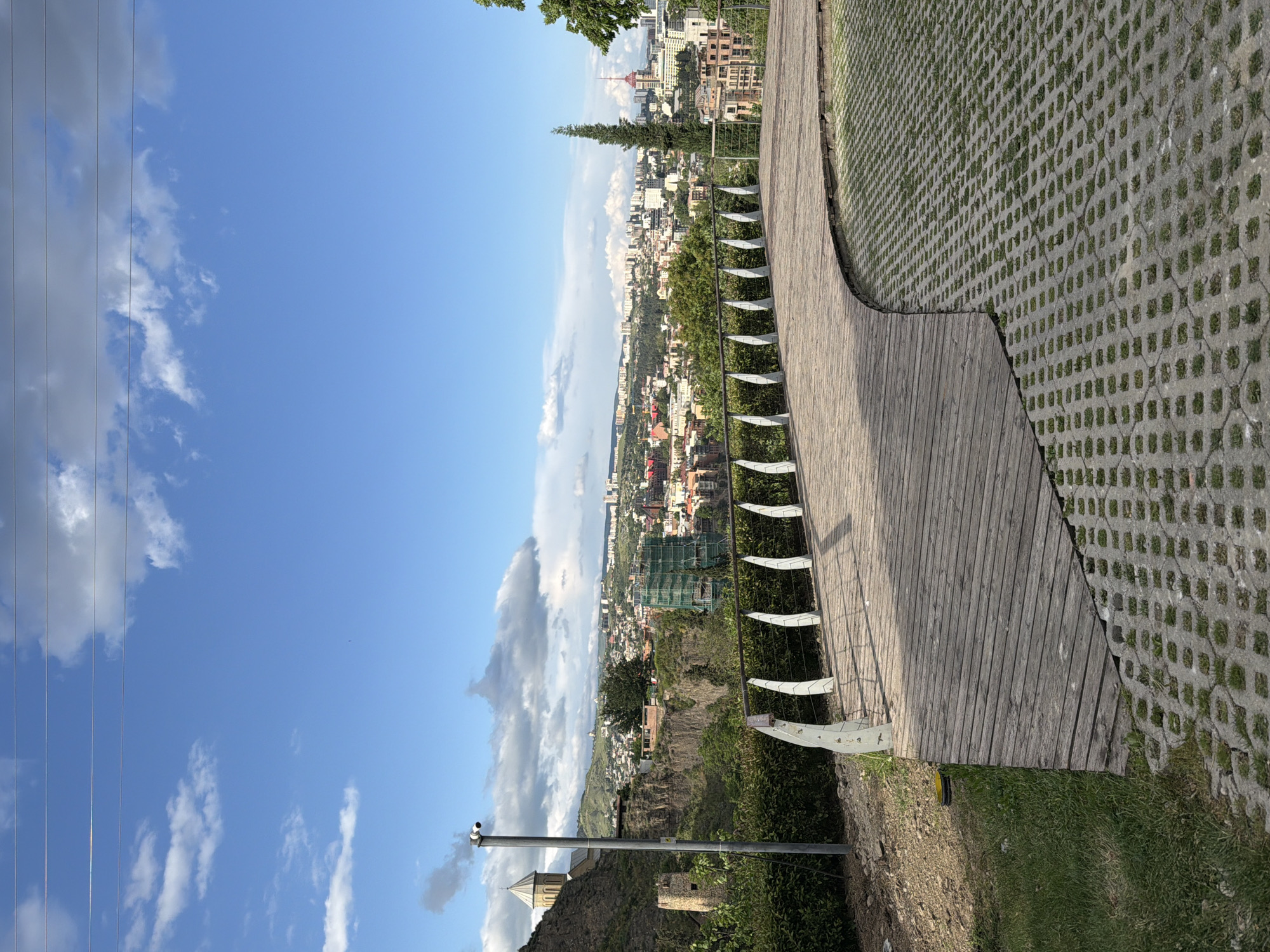 A bright, sunny day showcases the panoramic view of Tbilisi, Georgia, from a high vantage point. The foreground features a wooden walkway with a modern metal railing, extending alongside a patterned concrete and grass ground cover. Below, a cliffside reveals vegetation clinging to its slopes, with a historic stone tower and a security camera mounted on a pole at the bottom left. The cityscape of Tbilisi unfolds towards the horizon, marked by a mix of traditional and modern architecture, visible through the trees. The clear blue sky is adorned with scattered white clouds, completing the scenic backdrop.
