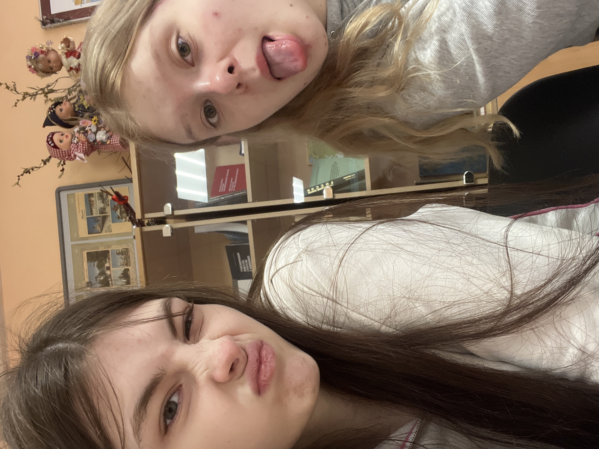 Two young women are taking a selfie indoors. The woman on the top, with blond hair, is sticking her tongue out. The woman on the bottom has dark brown hair, and is making a facial expression. In the background are bookshelves with books, picture frames, and decorative dolls. Based on the books and decor, the location might be a library or classroom in Saint Petersburg, Russia.
