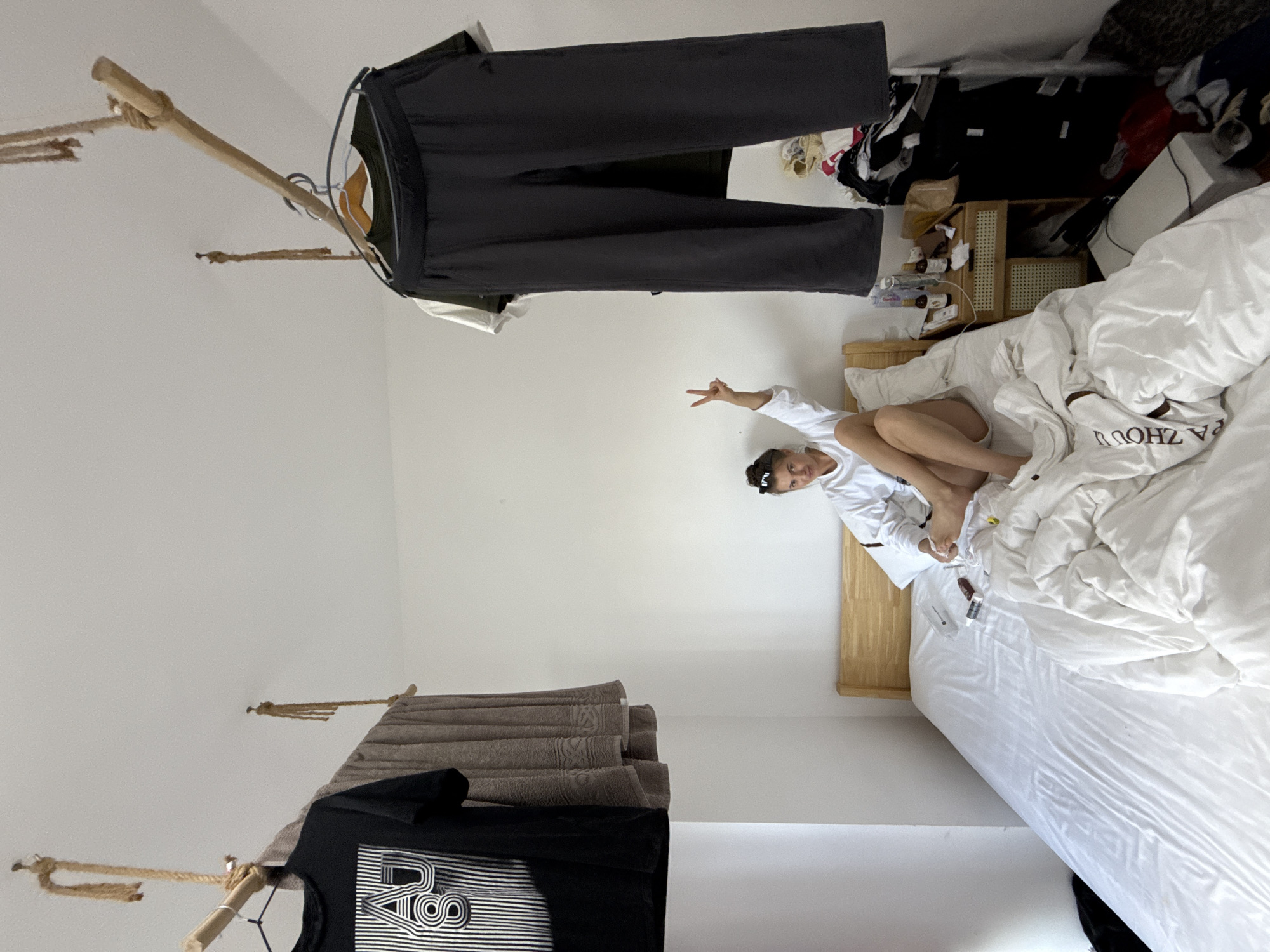 A woman is sitting on a bed in a bedroom with clothes hanging from ropes attached to the ceiling. The woman is wearing a white hoodie and dark shorts, and she is making a peace sign with her right hand. The bed is unmade and covered in white sheets. To the right of the bed is a nightstand with bottles and other items on it. In the background, there is a pile of clothes. The room appears to be in an apartment in Haizhu District, China.