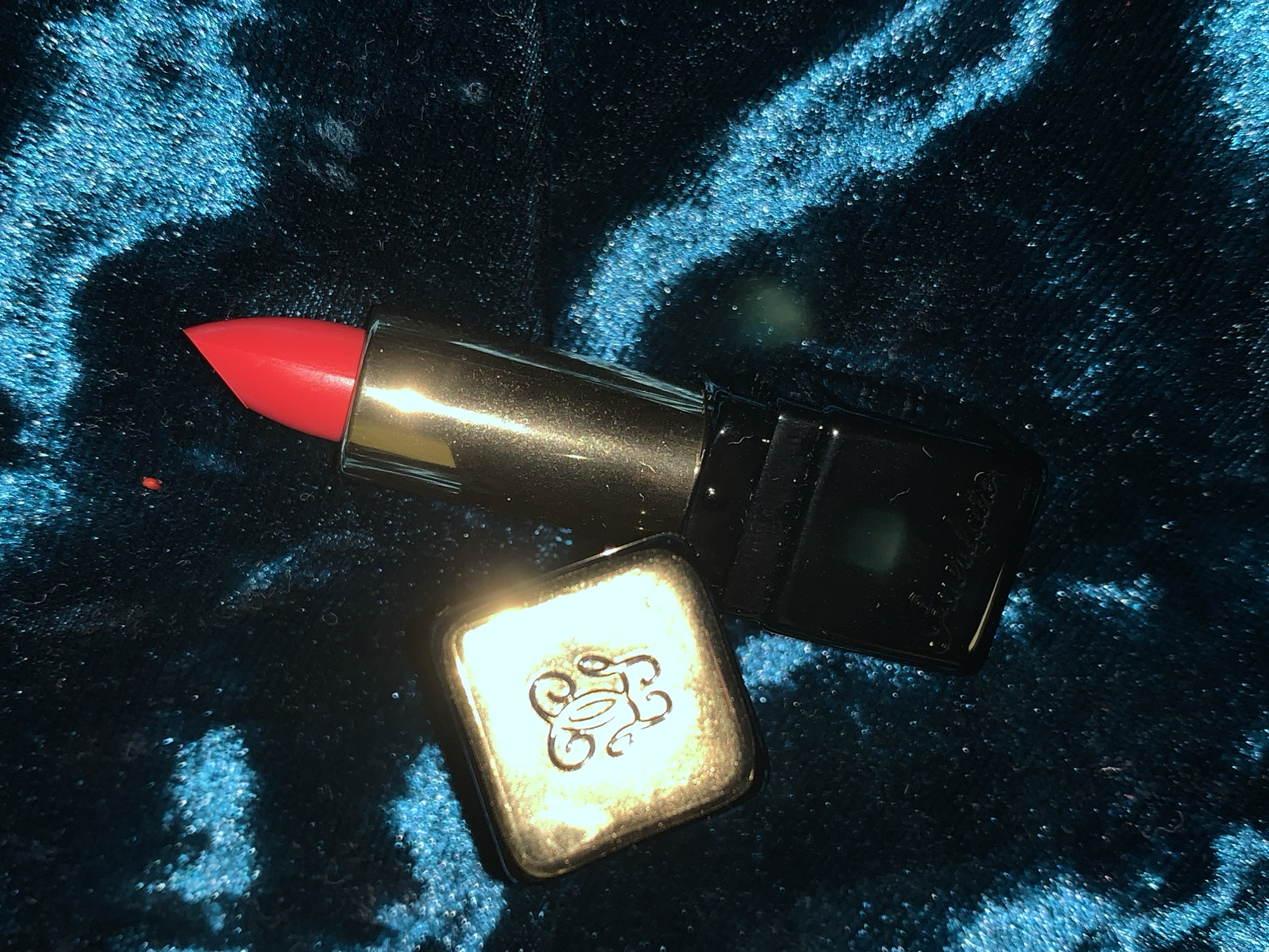 This image features a single, partially opened tube of lipstick resting on a dark teal, plush, textured fabric surface, likely velvet. The lipstick bullet, a vivid, uniform red, protrudes from a metallic gold component, which is attached to a dark, possibly black, rectangular base. The detached lipstick cap, a square-shaped metallic gold piece, is positioned to the left of the base. This cap displays a stylized, ornate double 'G' emblem on its upper surface, illuminated by a strong, localized light source, creating a bright highlight. The background fabric exhibits varying light reflections, resulting in luminous, lighter blue areas interspersed with darker, shadowed regions, emphasizing its soft texture. No individuals are present in the frame. The location is unknown.