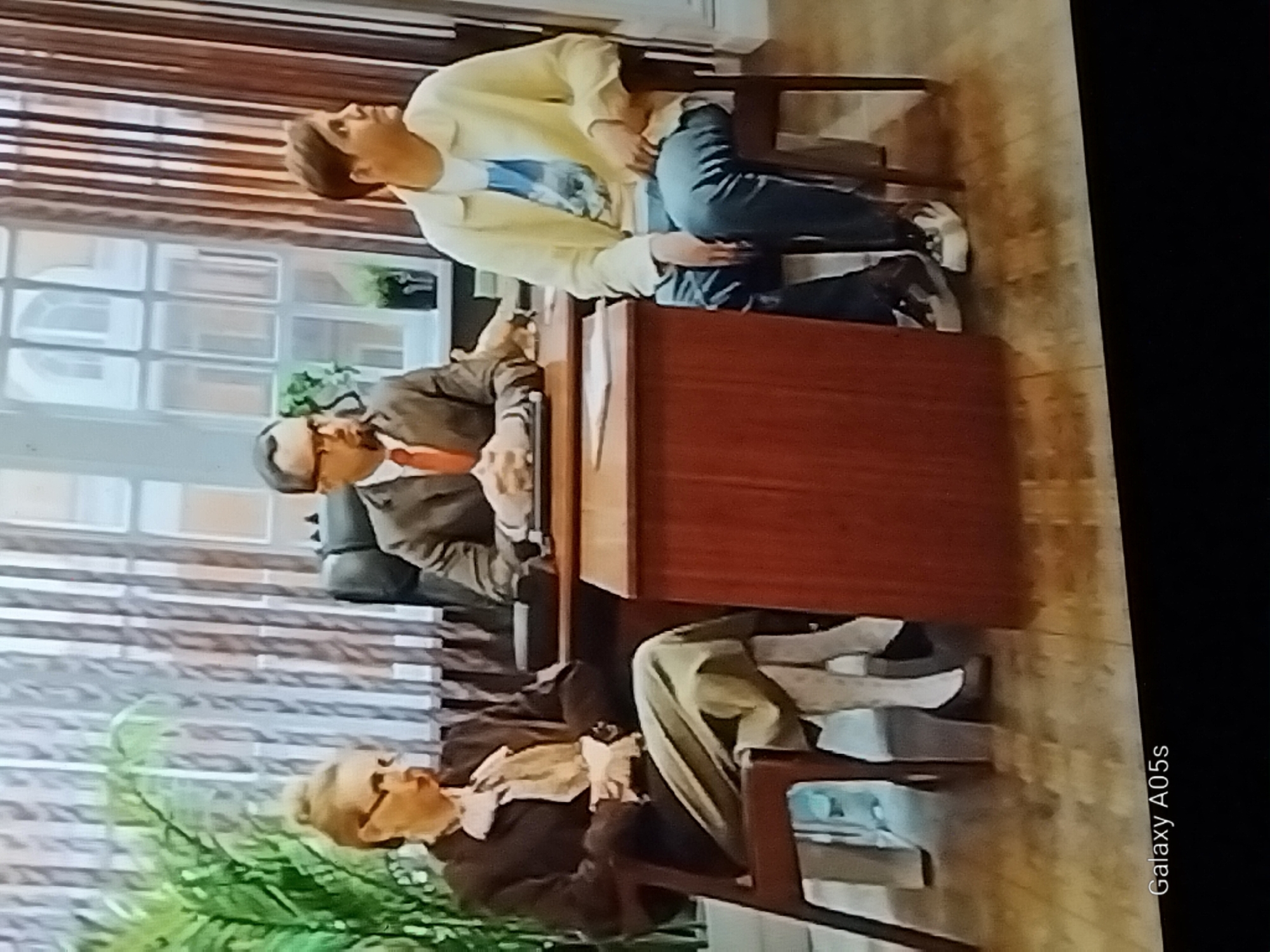The image shows three individuals seated in an office setting. On the left, a woman with blonde hair pulled back, wearing glasses, a dark jacket, a light-colored scarf, and a green skirt over patterned tights, sits in a dark wooden chair. In the center, a man with short dark hair, wearing glasses, a brown suit, and a red tie, sits behind a wooden desk. On the right, a person with short brown hair, wearing a light yellow jacket over a blue t-shirt and blue jeans, sits in a dark wooden chair. The room features vertical blinds in the background, a large window with architectural details, and a potted plant. The individuals appear to be engaged in a meeting or interview. There is no indication of the city of Druzhba, Russia in the image.