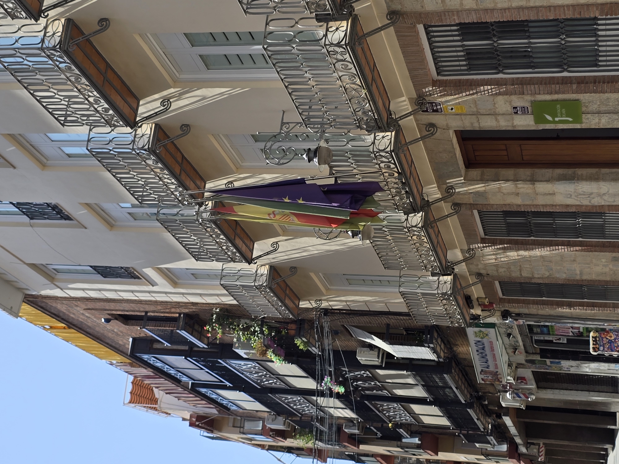 The image shows a street view in the city of Jaén, Spain, focusing on the facade of a building with multiple balconies and shops at the ground level. The building is light-colored with decorative wrought-iron railings on the balconies. Three flags are hanging from one of the balconies, with the Spanish flag and a purple flag with stars visible.
Below the balconies, there are shops with barred windows and doors. One shop has a sign that reads "ALI ALIMENTACION". A barber pole is visible outside one of the shops. Electrical wires and air conditioning units are also present on the building's facade. The street appears to be paved, and there is a bright blue sky overhead, suggesting it is a sunny day. The architecture suggests an urban environment.