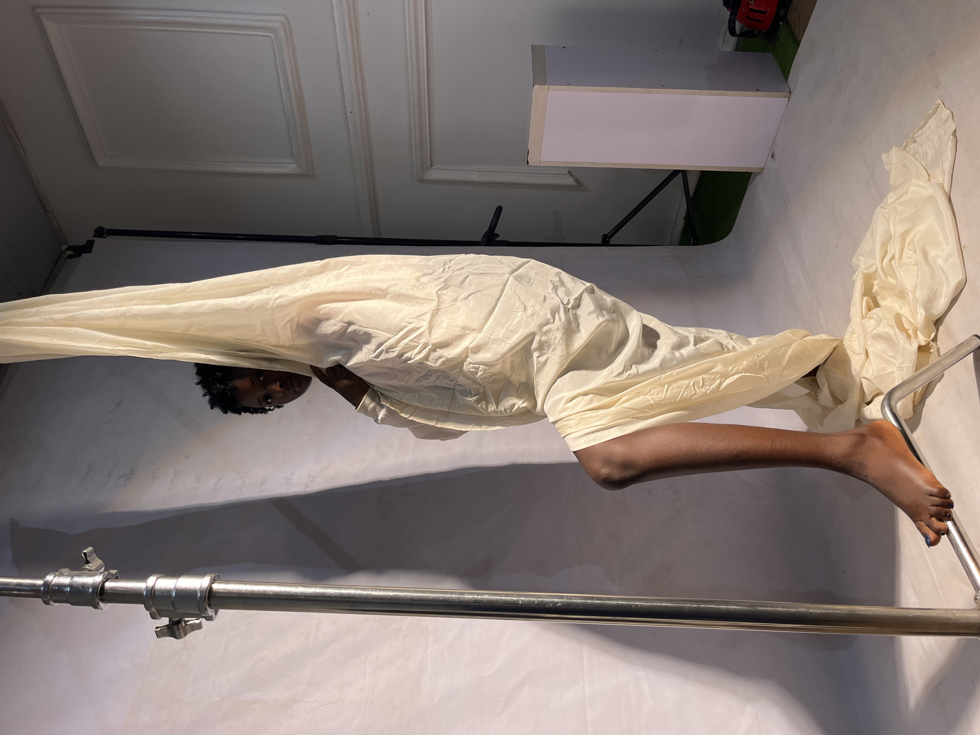 A person stands in a studio, partially obscured by a flowing, cream-colored fabric. They are positioned to the right of a tall, silver metal stand. Their left leg is bent and angled forward, with their foot resting on a horizontal metal bar. The fabric drapes around them, pooling on the floor at the bottom. The background consists of a white backdrop with some textured molding, and to the right, a small section of green turf is visible. A red object is partially visible in the far right background. The lighting casts shadows on the backdrop.