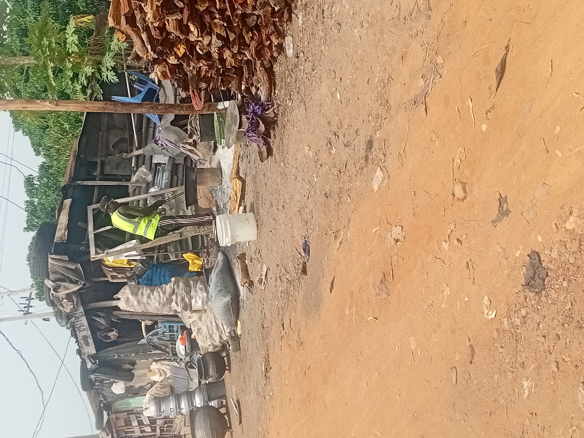 The image captures an outdoor, sunlit scene, likely a bustling roadside market or informal stall in Lokoja, Nigeria. The right side of the frame is dominated by a dusty, reddish-brown unpaved ground, scattered with small stones, dried leaves, and various pieces of debris like plastic fragments.
On the left, a cluttered, makeshift structure made of wooden planks and beams forms the core of the stall. A prominent feature at the top is a large pile of dried red items, resembling peppers or other produce, hanging from and resting on the structure. Below this, a blue plastic chair is propped on a wooden beam.
Various items are arranged or stored around the stall: white plastic buckets, several metal pots or containers, bundles of fabric or sacks (including grey, blue, and yellow ones), and a large, grey-covered object that could be a fish or another wrapped commodity. Further to the left, parts of a vehicle, possibly a truck or bus, are visible, including tires and what appears to be a steering wheel. Green foliage from trees is visible in the background on the far left, and faint utility wires cross the upper left corner against a bright, clear sky.
A single individual, a dark-skinned person, is visible in the mid-left portion of the image. They are wearing a bright yellow or lime green reflective vest over a dark top and striped pants, and appear to be leaning over or engaged in some activity within the stall area. Their face is not clearly visible, so no specific emotions can be discerned. The scene suggests a busy commercial day, characteristic of an open-air market. No legible text is visible in the image.