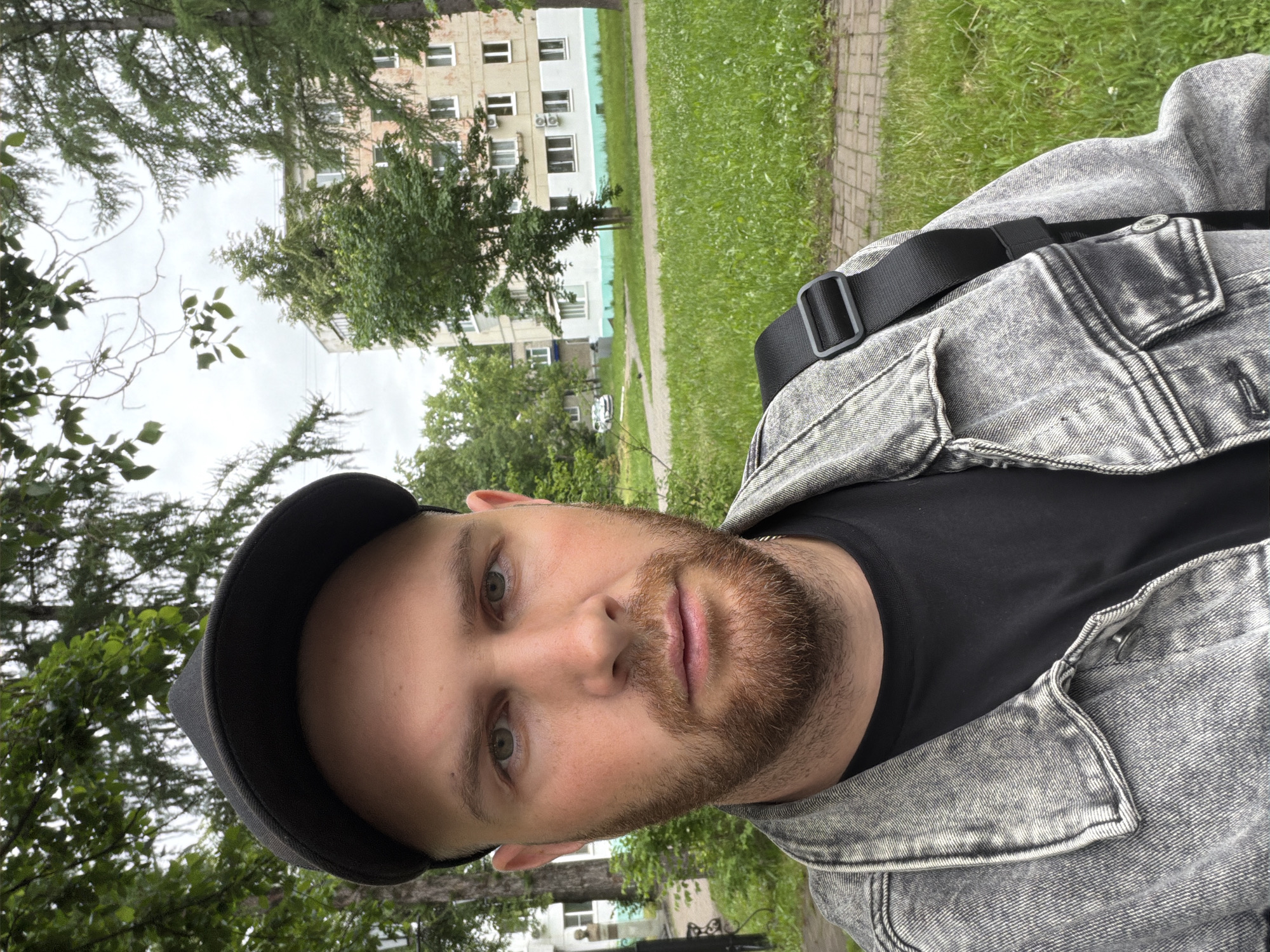 A male individual is depicted in a close-up selfie-style photograph, standing outdoors in what appears to be an urban park area within the city of Chaikovsky, Russia, under an overcast sky. The subject, a man in his late 20s to early 30s, possesses a light skin tone, a neatly trimmed brown beard, and light green eyes. He maintains a neutral expression and looks directly at the camera. He wears a black baseball cap with the brim facing forward, a faded grey acid-wash denim jacket, and a plain black t-shirt underneath. A black textile strap from a shoulder bag is visible over his right shoulder.
The background consists of dense green foliage, including various deciduous trees and coniferous trees with needle-like leaves, indicative of late spring or summer. To the immediate left of the subject, partially obscured, is a small, dark-colored bust of a male figure atop a light-colored rectangular pedestal, situated next to a tree and a paved path. Further in the background, to the left, a light-colored multi-story building is partially visible. To the right of the subject, a beige or pale orange multi-story apartment building with numerous white-framed windows and balconies is prominent. The lower section of this building is painted white with a distinctive pale turquoise-green band above it. Several external air conditioning units are mounted below the windows. The ground in the foreground and midground features patches of green grass and segments of light-colored interlocking paving stones and asphalt paths.