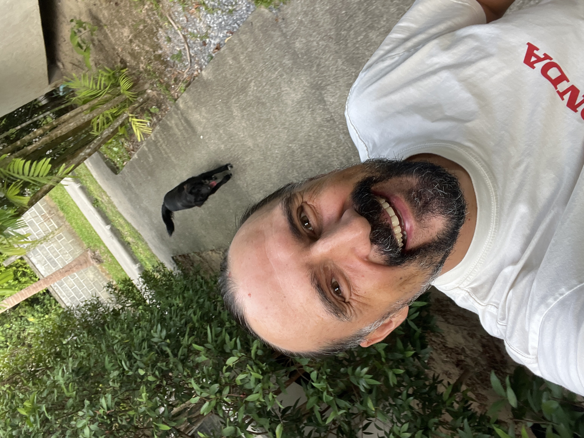 The image appears to be a selfie taken by a man with a black dog visible in the background. The man has a dark beard with some gray hairs and is wearing a white t-shirt with the word "HONDA" printed on it in red. He is smiling. The background includes a concrete walkway or driveway, gravel, and various types of greenery including some palm trees. Based on the prompt, this scene is located in บ้านบางรักษ์, Thailand.
