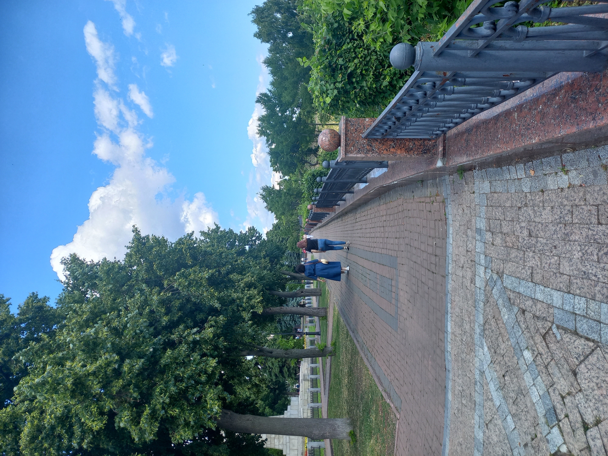 The image shows a paved walkway in a park in Ulyanovsk, Russia, with a row of trees on the left and a metal railing with a stone embankment on the right. Two women are walking away from the camera on the walkway. The sky is blue with scattered white clouds. In the background, there is dense green foliage. The walkway is made of red brick pavers, with a decorative pattern of grey and white stone in the foreground. The railing is black metal with ornate scrollwork, and the embankment is made of reddish-brown granite. The trees are tall and leafy, providing shade to the park. The scene is brightly lit, suggesting it is daytime.