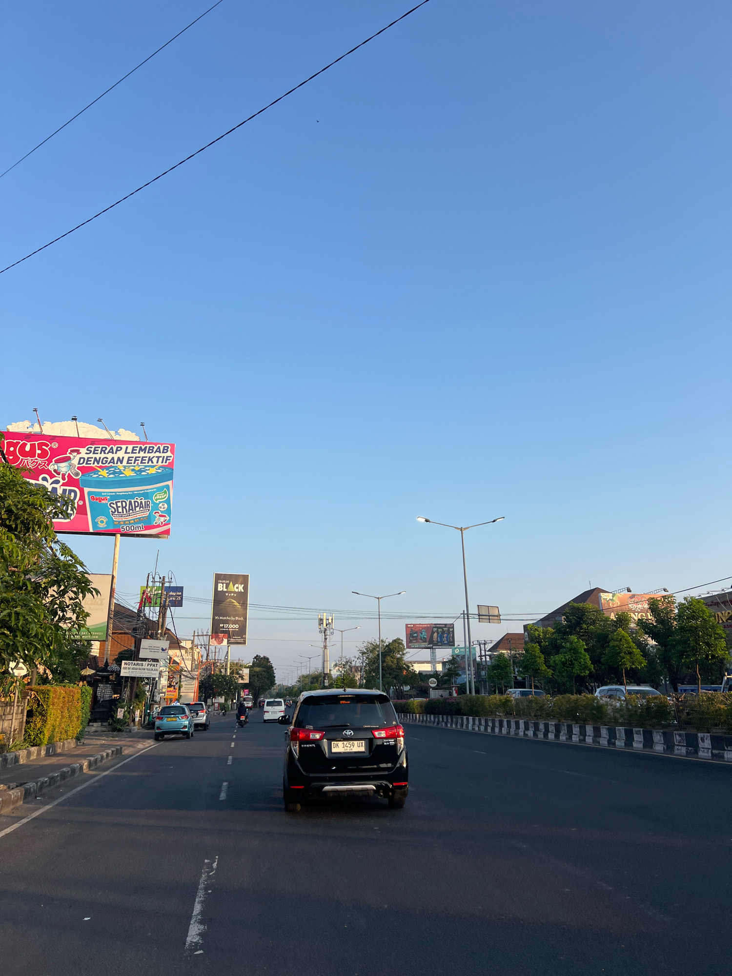 The image depicts a view of a street in Kuta, Indonesia, likely in the late afternoon or early morning based on the lighting. A black SUV is the most prominent vehicle in the foreground, driving down a multi-lane road. To the left of the road, there is a sidewalk lined with trees and bushes, and a series of billboards. One large billboard on the left advertises "Bagus SERAPAIR" with images of water and a product container. Another billboard further down the street shows "BLACK MATCHA". Buildings and more trees are visible on the right side of the road, along with streetlights. The sky is clear and blue, with two thin power lines running diagonally across the upper portion of the frame. Several other vehicles, including cars and motorcycles, are visible further down the road, indicating moderate traffic. The general environment suggests a commercial or residential area within the city.