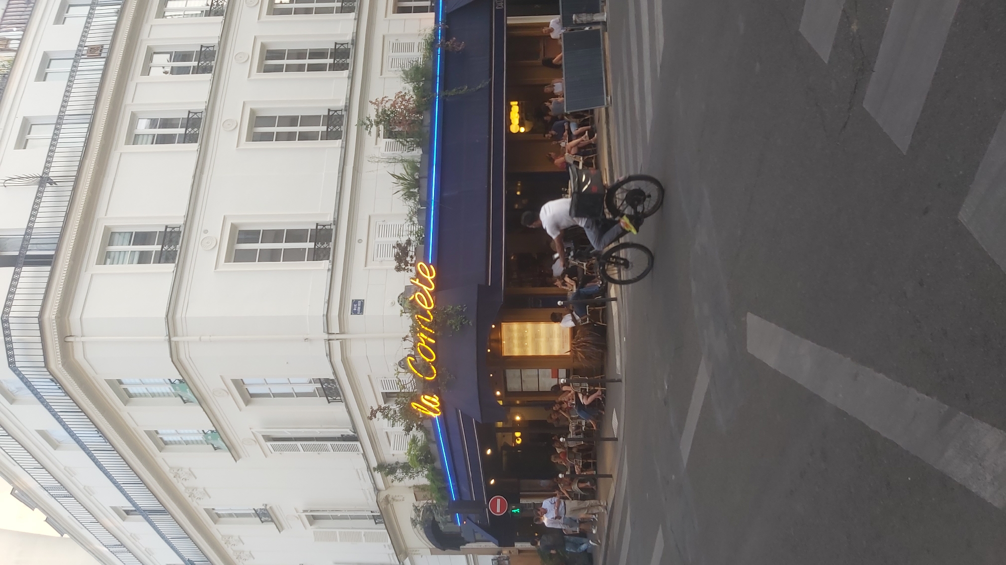 An eye-level, full shot captures a street scene in Paris, France, on a bright day. The main focus is a corner building with a restaurant, "La Comète," occupying the ground floor. The restaurant has a navy blue awning and the name "La Comète" is displayed in bright yellow neon lettering. Outside the restaurant, tables and chairs are occupied by patrons. A man on a bicycle, wearing a white t-shirt and dark pants, is in the foreground, riding across a pedestrian crossing. He is wearing a black bag on the back of his bicycle. The street is paved with asphalt, and there are white crosswalk markings. The building itself is white, with multiple floors, featuring numerous windows with black frames and some with shutters. A black metal railing adorns a balcony on the upper floors. A street sign is visible on the corner of the building. The overall impression is a lively urban environment.