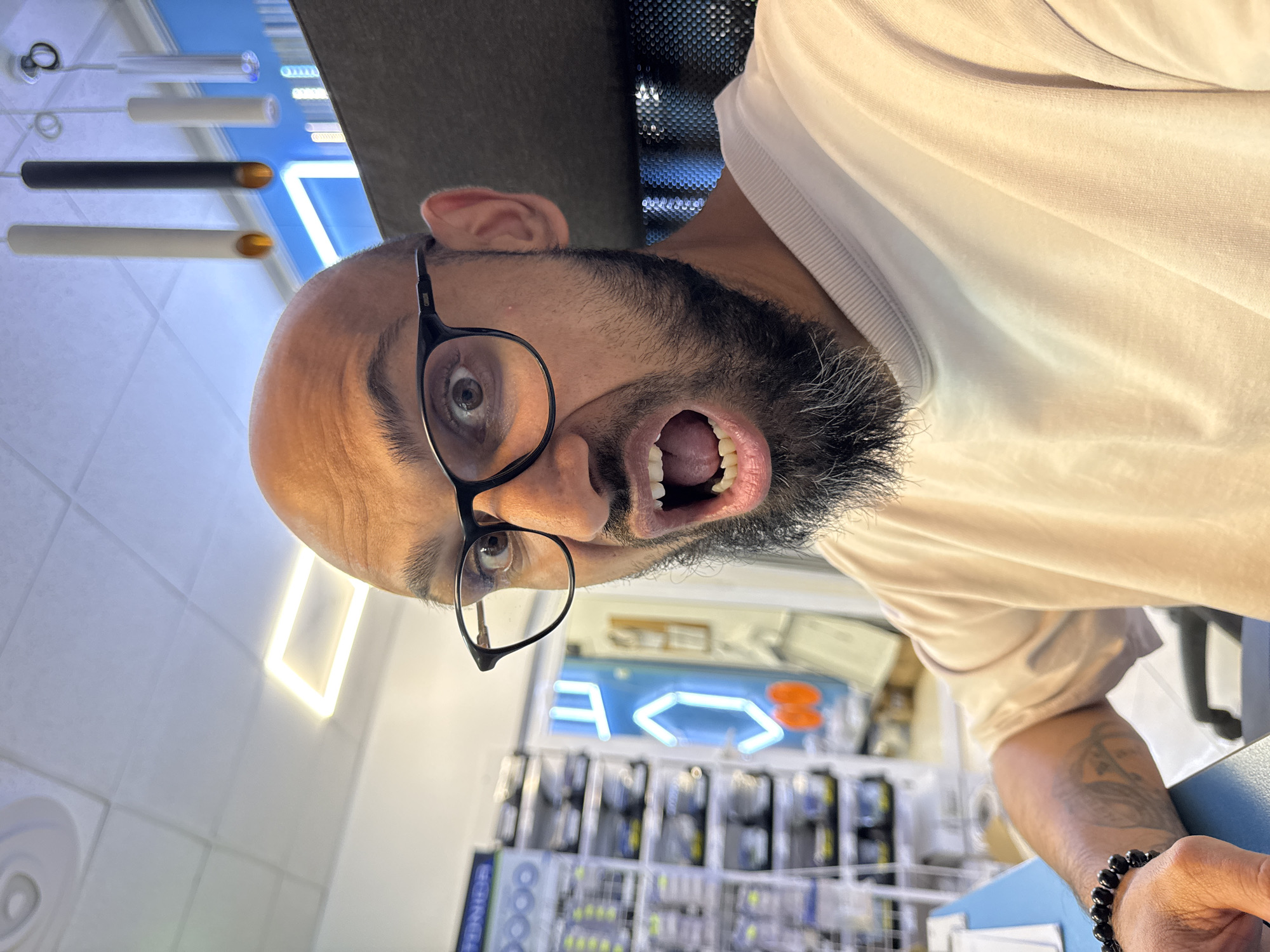 A close-up, waist-up portrait depicts a bald man with dark-rimmed glasses and a dark beard exhibiting graying strands, seated at a desk in a commercial or office environment located in Plovdiv, Bulgaria. The subject directly faces the camera with wide-open eyes and an agape mouth, revealing teeth and tongue, indicative of an expressive, possibly startled or shouting, action. A black beaded bracelet is visible on the subject's left wrist, and a tattoo is partially discernible on the left forearm, beneath the sleeve of a light-colored, crew-neck t-shirt. The background on the left features shelving units stocked with numerous small, packaged items. A prominent blue wall section in the mid-ground is adorned with a neon sign displaying abstract shapes, including an inverted 'E' or 'F', a hexagonal form, and an orange component. The ceiling above consists of white acoustic tiles, integrated with a square LED light fixture and several modern cylindrical pendant lights in white and black, some emitting an amber glow. A dark mesh-backed office chair is partially visible behind the subject on the right.