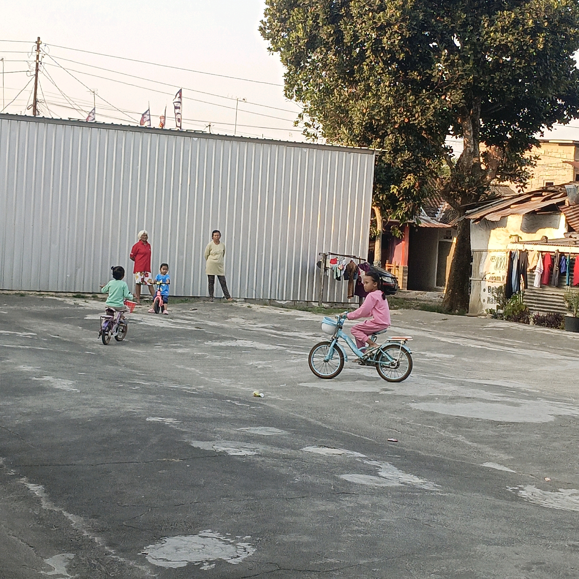 The image depicts an outdoor scene in Salatiga, Indonesia. A group of people are present, including at least two children riding bicycles and two adults standing. One child, wearing a light blue outfit, is riding a small pink bicycle. Another child, in a mint green outfit, is on a purple bicycle, facing away from the camera. A woman in a red shirt and patterned skirt and another woman in a light yellow outfit are standing in the background. The children appear to be in a paved outdoor area, possibly a courtyard or street, with a large, corrugated metal building behind them. A mature tree with dense foliage is visible to the right of the building. Adjacent to the building, there are houses with corrugated tin roofs. Clothes are hanging on a clothesline between the metal building and the houses. The ground is paved and shows signs of wear and some light-colored stains. The sky is clear and appears to be late afternoon or early evening, with the setting sun casting a warm glow on the scene.