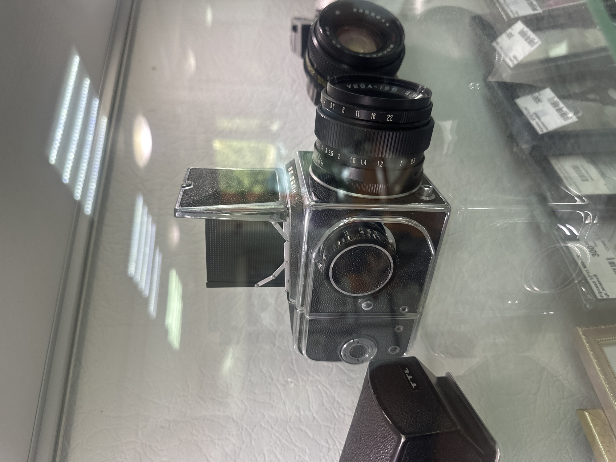 The image shows a Hasselblad 88 camera on display in a store. The camera is black and silver with a lens attached. There is also a brown leather case for the camera next to it. The camera is on a glass shelf, and reflections of fluorescent lights are visible on the shelf and the camera. In the background, there are other items on display, including what appear to be price tags. The scene is indoors, likely in a retail environment. The metadata indicates the location is Tobolsk, Russia. There are no people visible in the image, and no actions are being performed.