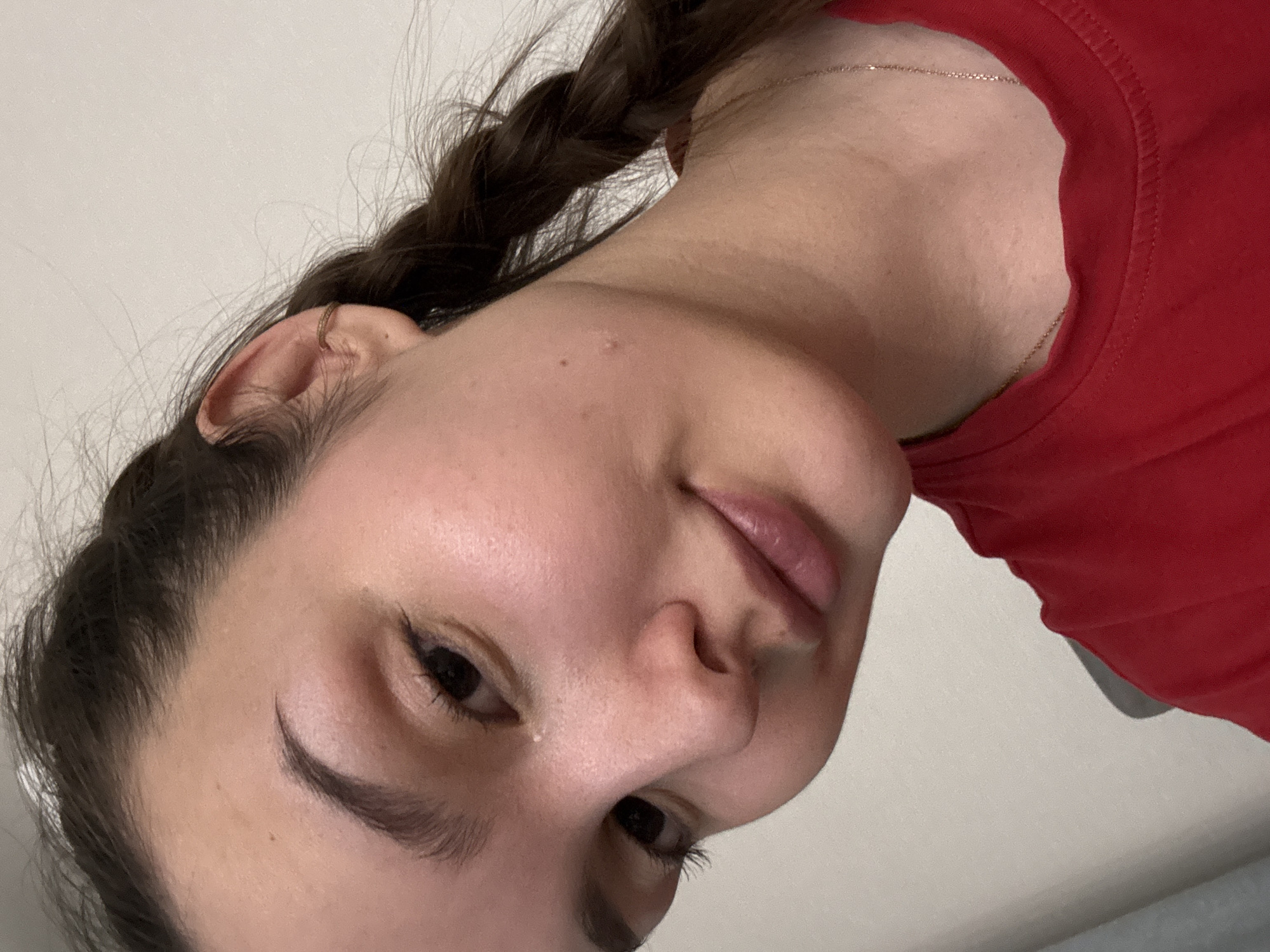 A close-up, tilted photograph shows a woman with dark hair in a braid and a red shirt. A thin gold necklace is visible on her neck. Her face is clear, with minimal makeup. The location cannot be determined from the image.