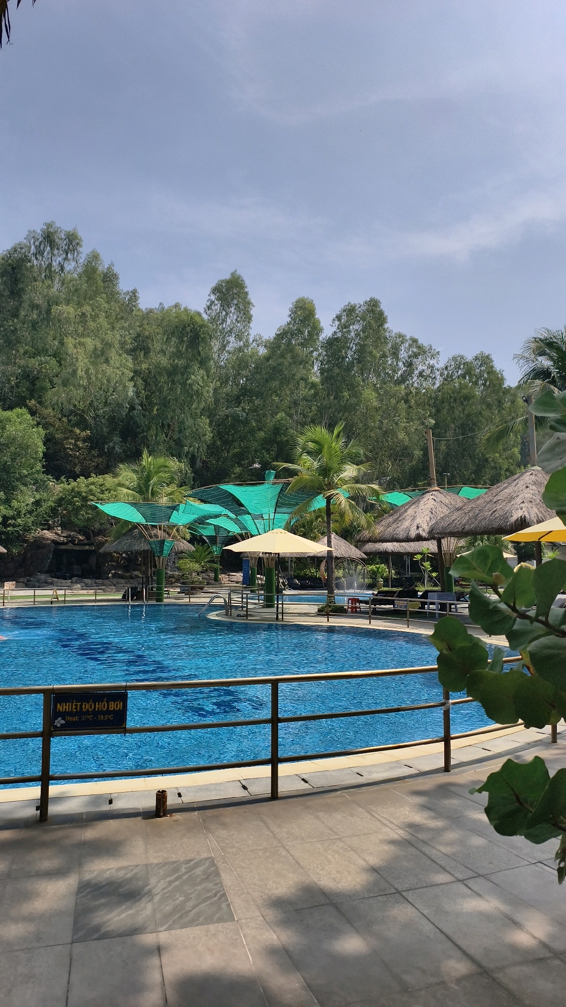 The image shows a swimming pool area, likely at a resort or hotel, in Nha Trang, Vietnam. The pool is large and blue, with a textured bottom. Around the pool, there are lounge chairs, umbrellas with thatched roofs, and some covered seating areas with green netting. Palm trees and other lush green foliage are visible in the background, with a dense line of trees and hills beyond. The sky is clear with some wispy clouds. A sign near the edge of the pool reads "NHIỆT ĐỘ HỒ BƠI" with smaller text below indicating temperature ranges. The foreground shows a tiled walkway and a metal railing. There are no people prominently featured in the image.