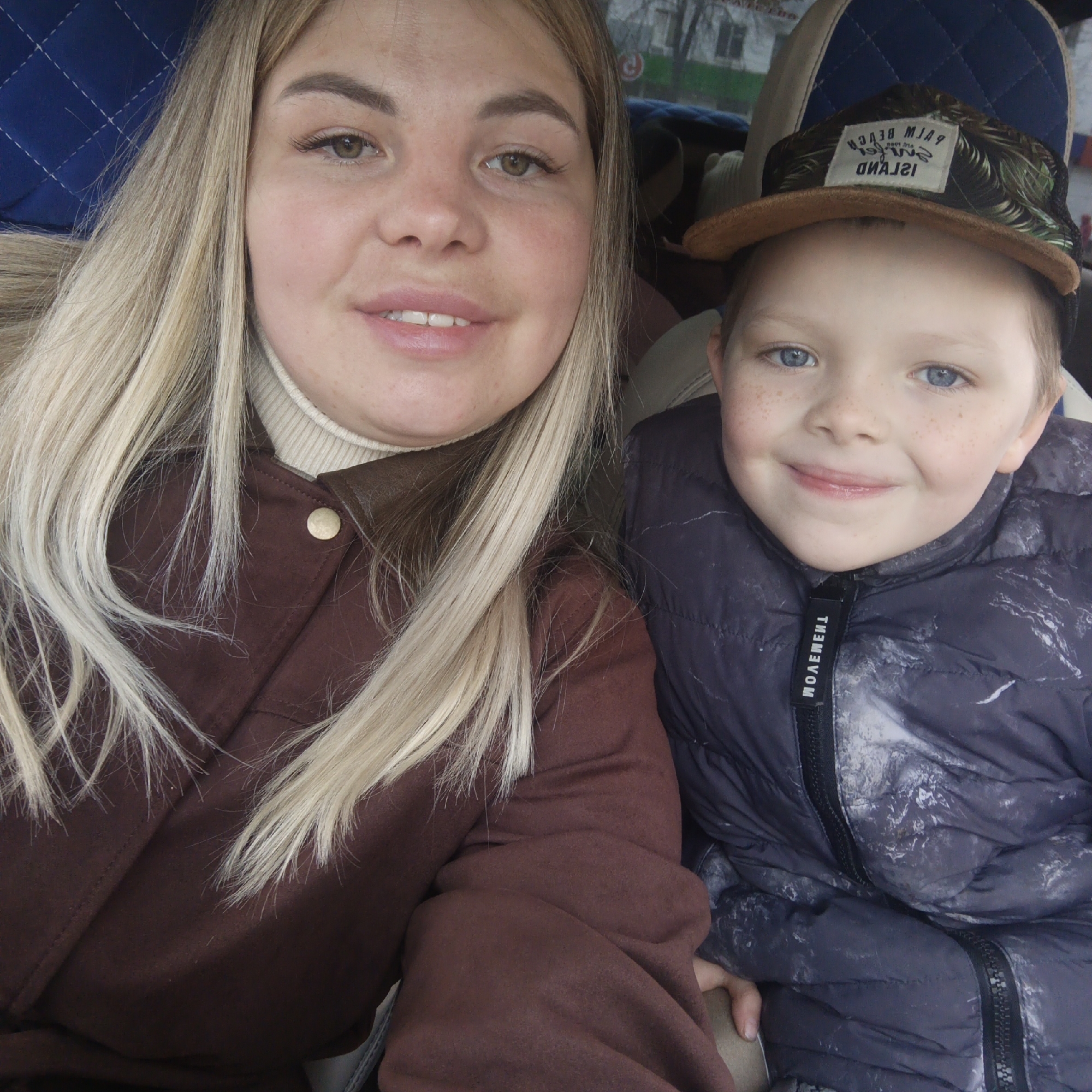 A woman and a young boy are indoors, likely in a vehicle, as suggested by the blue quilted seat visible. The woman has long blonde hair, fair skin, and wears a brown jacket over a light-colored turtleneck. The boy has light-blue eyes, freckles, and wears a patterned jacket with a black zipper and collar. The background is out of focus. The location is Unknown, Unknown.