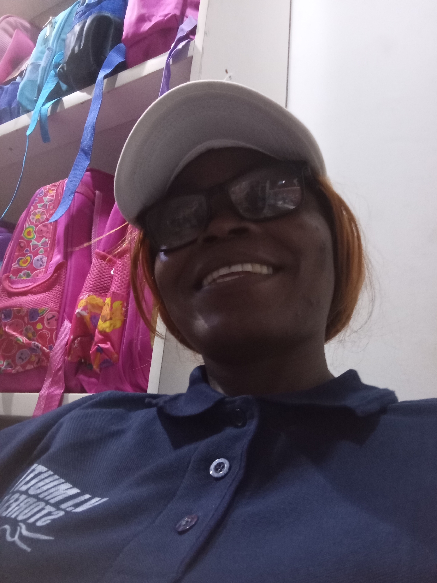 In a close-up, low-angle selfie, a Black woman with reddish-brown hair and glasses smiles at the camera. She is wearing a dark blue collared shirt and a white baseball cap. Behind her, on shelves, are several brightly colored backpacks, primarily pink and blue, suggesting a storage area or possibly a school environment. The background wall is plain white. The image appears to have been taken indoors, possibly in a residential setting or a place of business.