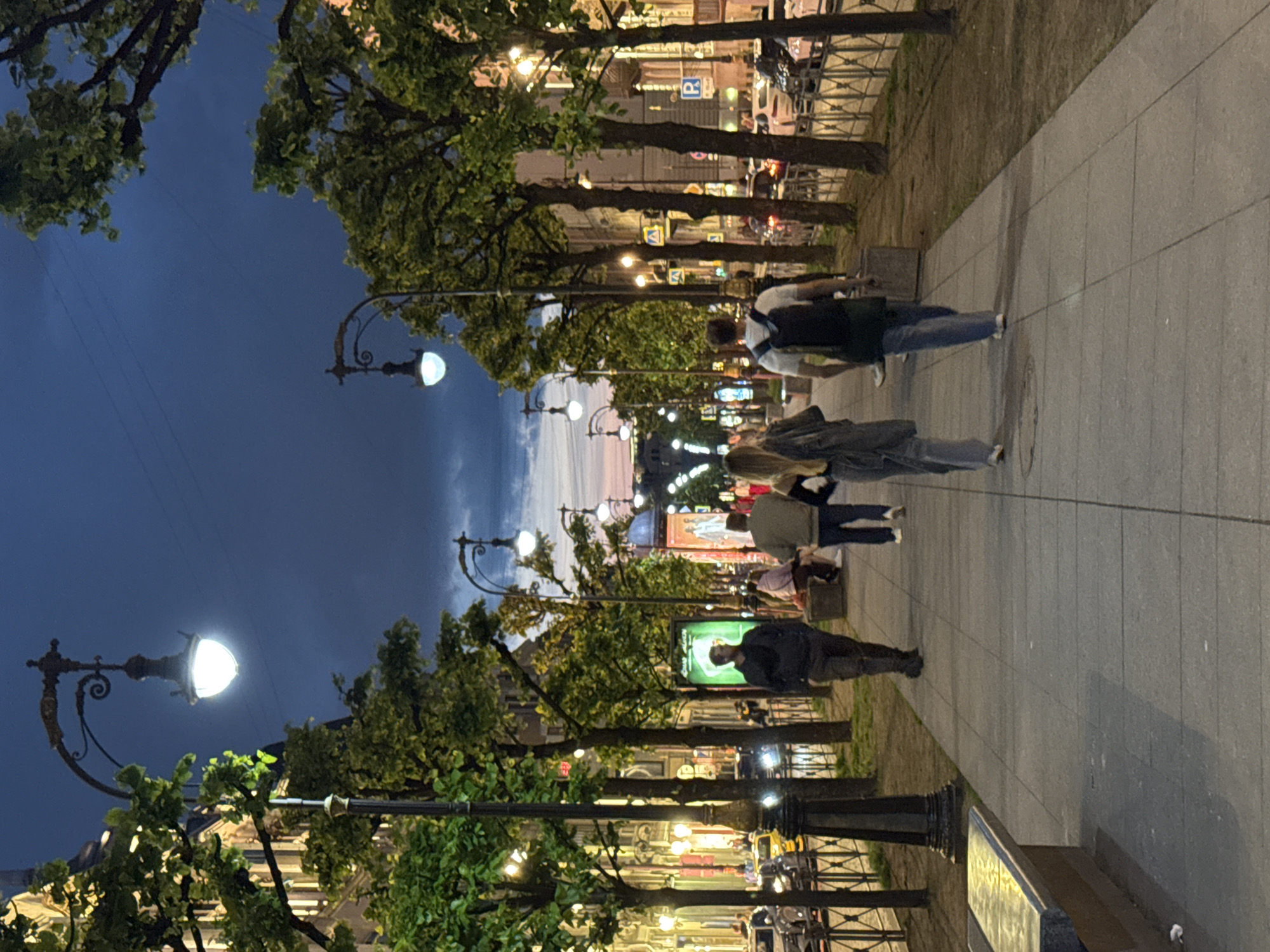 The image depicts Nevsky Prospekt in Saint Petersburg, Russia, at dusk. The wide pedestrian sidewalk is lined with mature trees, casting shadows, and ornate streetlights illuminate the scene. Several individuals are walking along the sidewalk. In the foreground, two people are walking away from the camera, one wearing a backpack. Further down the sidewalk, another person is walking towards the camera. On the left side of the sidewalk, a bench is visible. On the right side of the sidewalk, a street with cars and buildings can be seen. The sky is a deep blue with hints of pink from the setting sun, and clouds are present.
