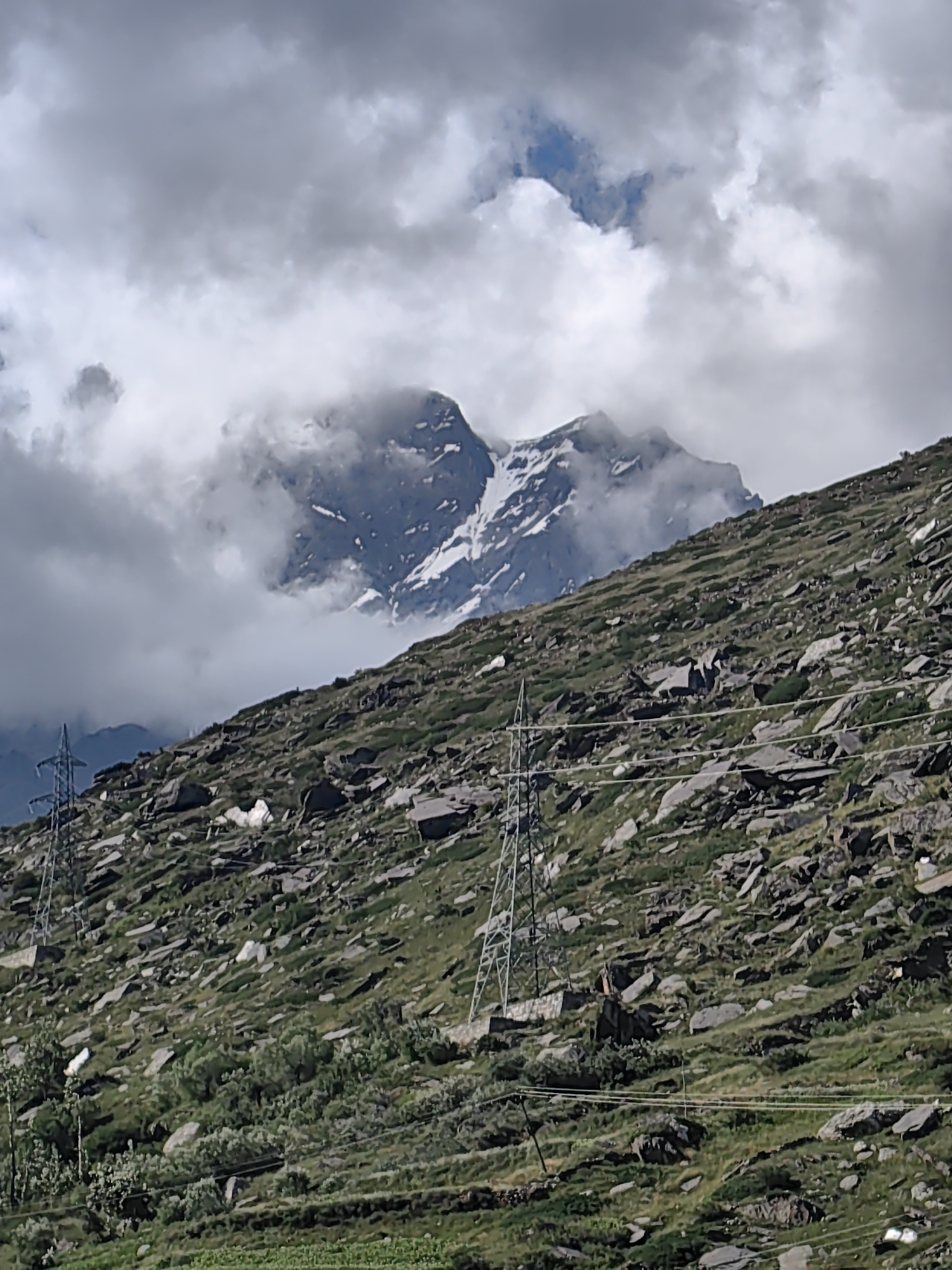The image depicts a mountainous landscape under a cloudy sky. A steep, rocky slope dominates the foreground, covered in sparse green vegetation and large stones. Two electricity pylons are visible on the left and right sides of the slope. In the background, a majestic mountain peak with snow-capped summits emerges from behind a curtain of clouds. The lighting suggests it is daytime, likely overcast.