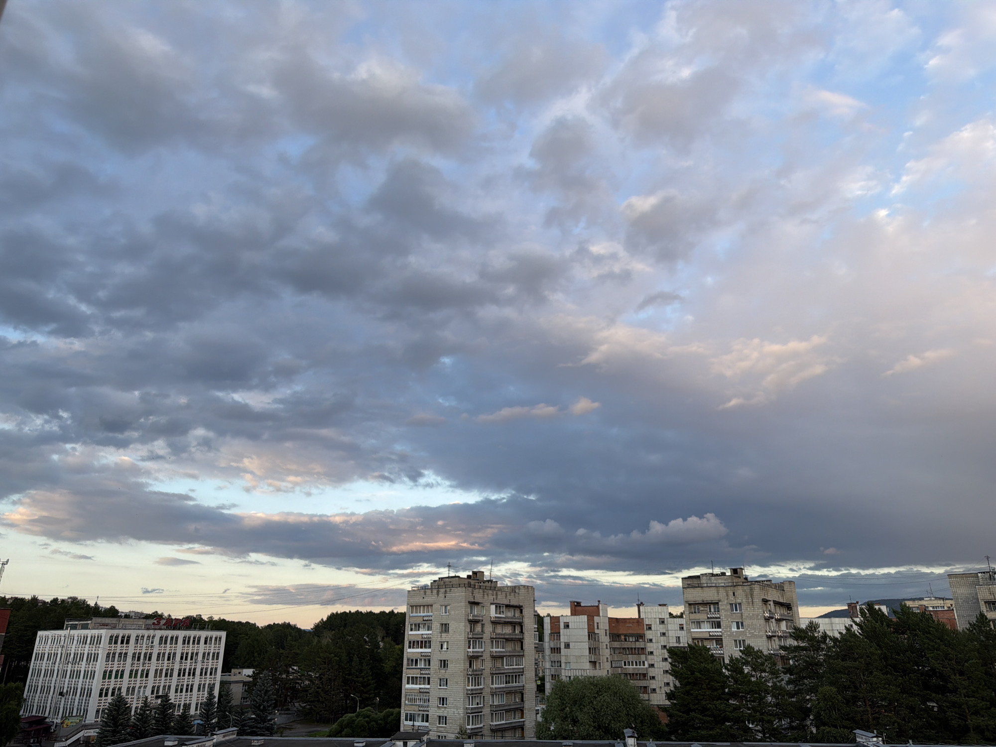 The image captures an urban landscape under a dramatic sky filled with a mix of dark, heavy clouds and lighter, brighter patches. The setting appears to be a city in Tryokhgorny, Russia, with several multi-story buildings and a dense line of trees forming a backdrop.
Dominating the lower portion of the image are several buildings. On the far left, a modern white building with a distinctive geometric facade stands out. To its right, a series of older, concrete high-rise apartment buildings are visible, some appearing weathered. These buildings have numerous windows and balconies, typical of Soviet-era architecture. A cluster of dark green pine trees partially obscures some of the buildings, adding a natural element to the urban scene.
The sky is a significant feature, with a complex cloud formation suggesting dynamic weather conditions. The clouds range in color from deep gray to soft white, with hints of pink and orange appearing where the light breaks through. This suggests either a sunrise or sunset, with the time of day likely being early morning or late evening. The weather appears overcast with the possibility of rain.
There is some visible text on the top of the white modern building, appearing as red letters. The overall mood conveyed is one of an urban environment transitioning through the day, with a strong emphasis on the powerful and ever-changing sky. The scene is devoid of people, focusing solely on the architecture and the natural elements.